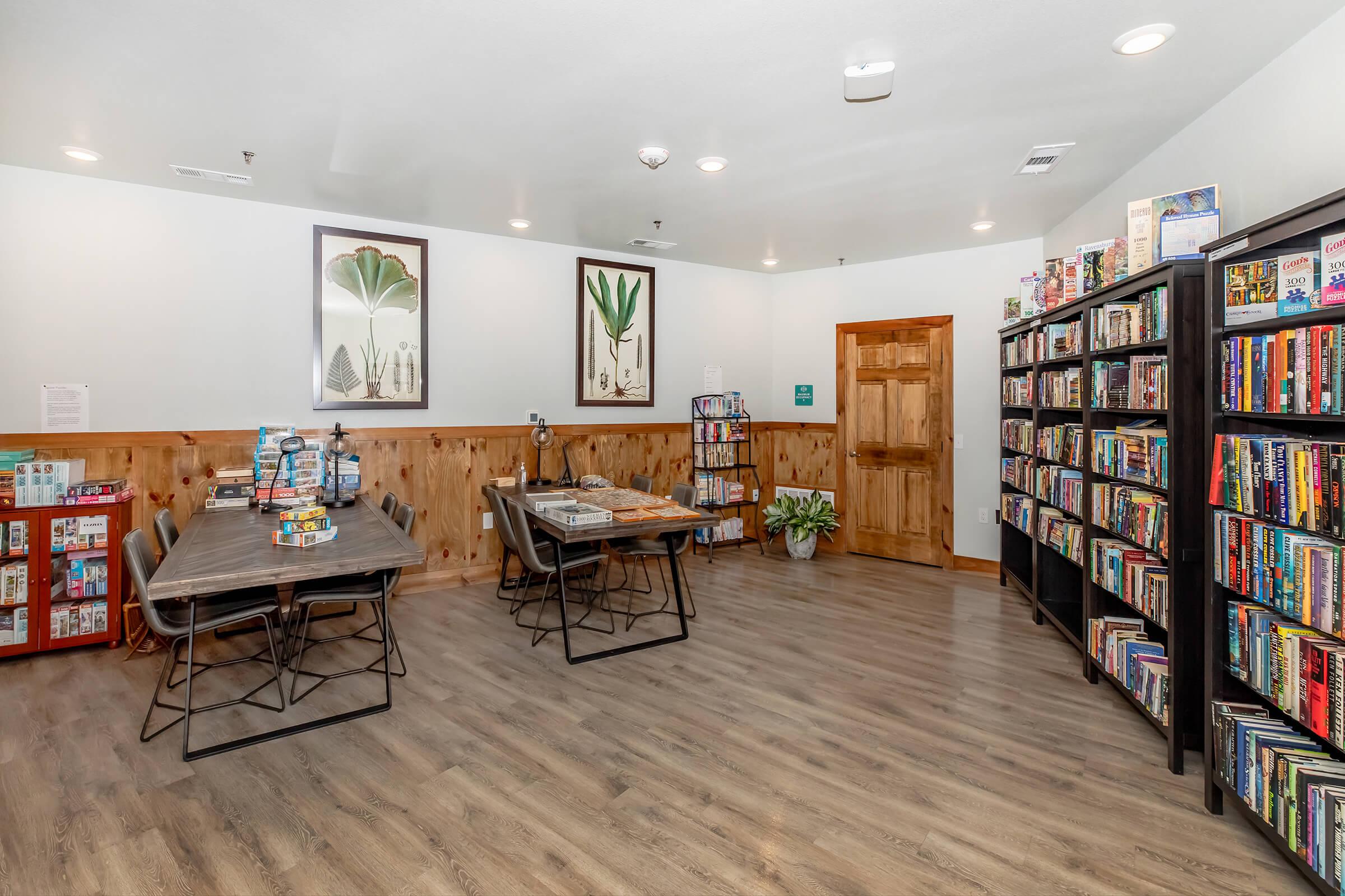 A cozy room featuring a wooden floor, shelves filled with books, a table with board games, and decorative plants. The walls showcase framed botanical prints and there is warm lighting, creating a welcoming environment for reading or playing games.