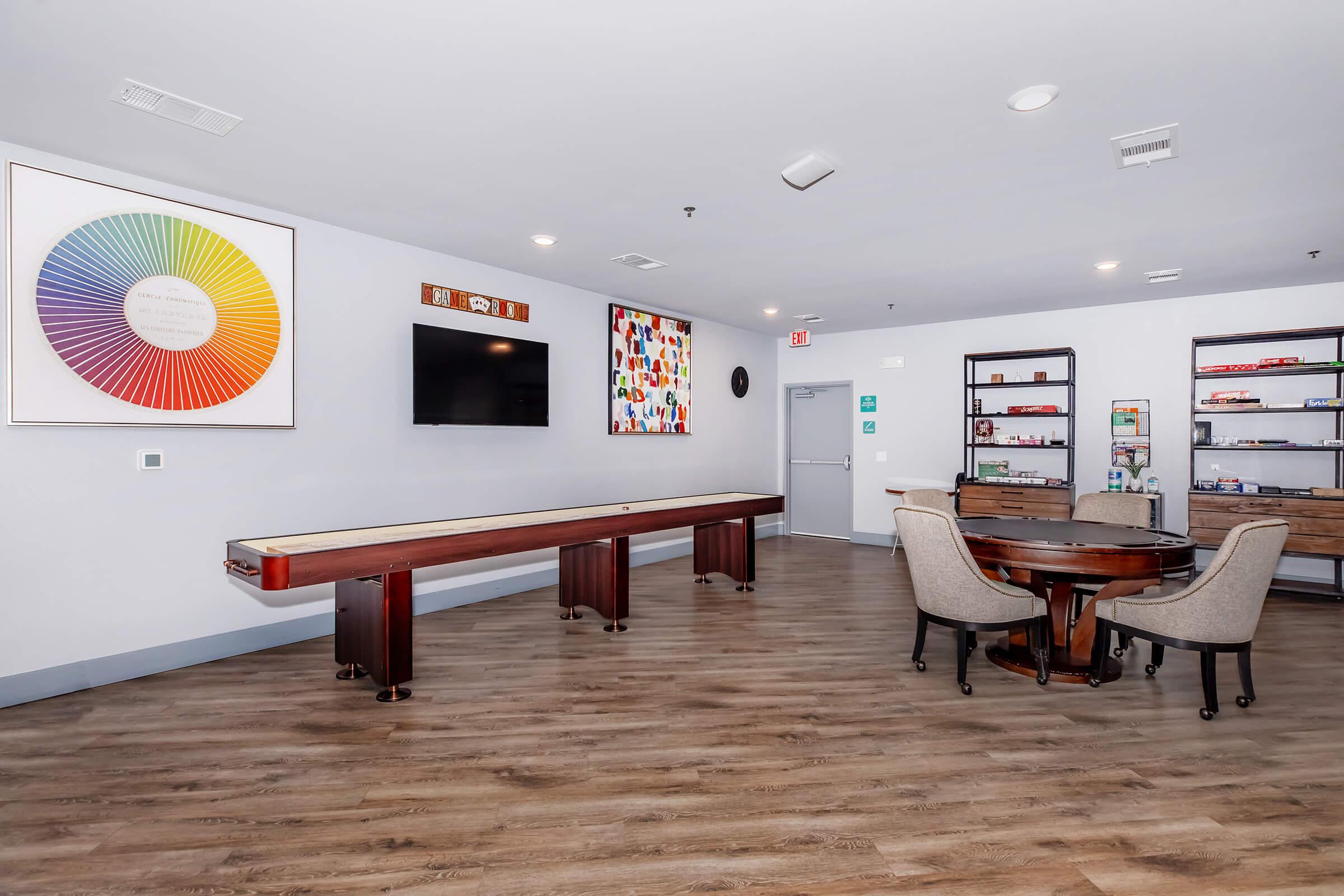 A spacious room featuring a shuffleboard table along one side, a round dining table with upholstered chairs, a wall-mounted TV, and colorful artwork. The decor includes a large rainbow color wheel and various shelves displaying items, all set against a light-colored wall and wooden flooring.