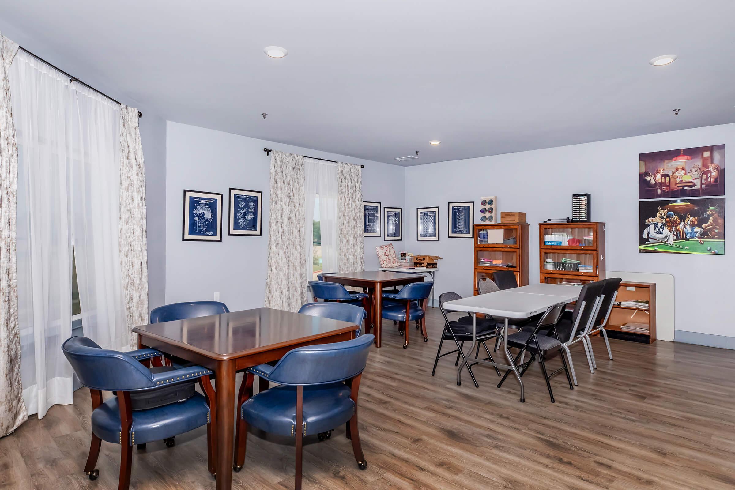 A well-lit common room featuring several tables with chairs, light wooden flooring, and large windows adorned with sheer curtains. The walls are decorated with framed pictures, and there is a wooden shelf with games and books. The room has a welcoming and spacious atmosphere, suitable for social gatherings.