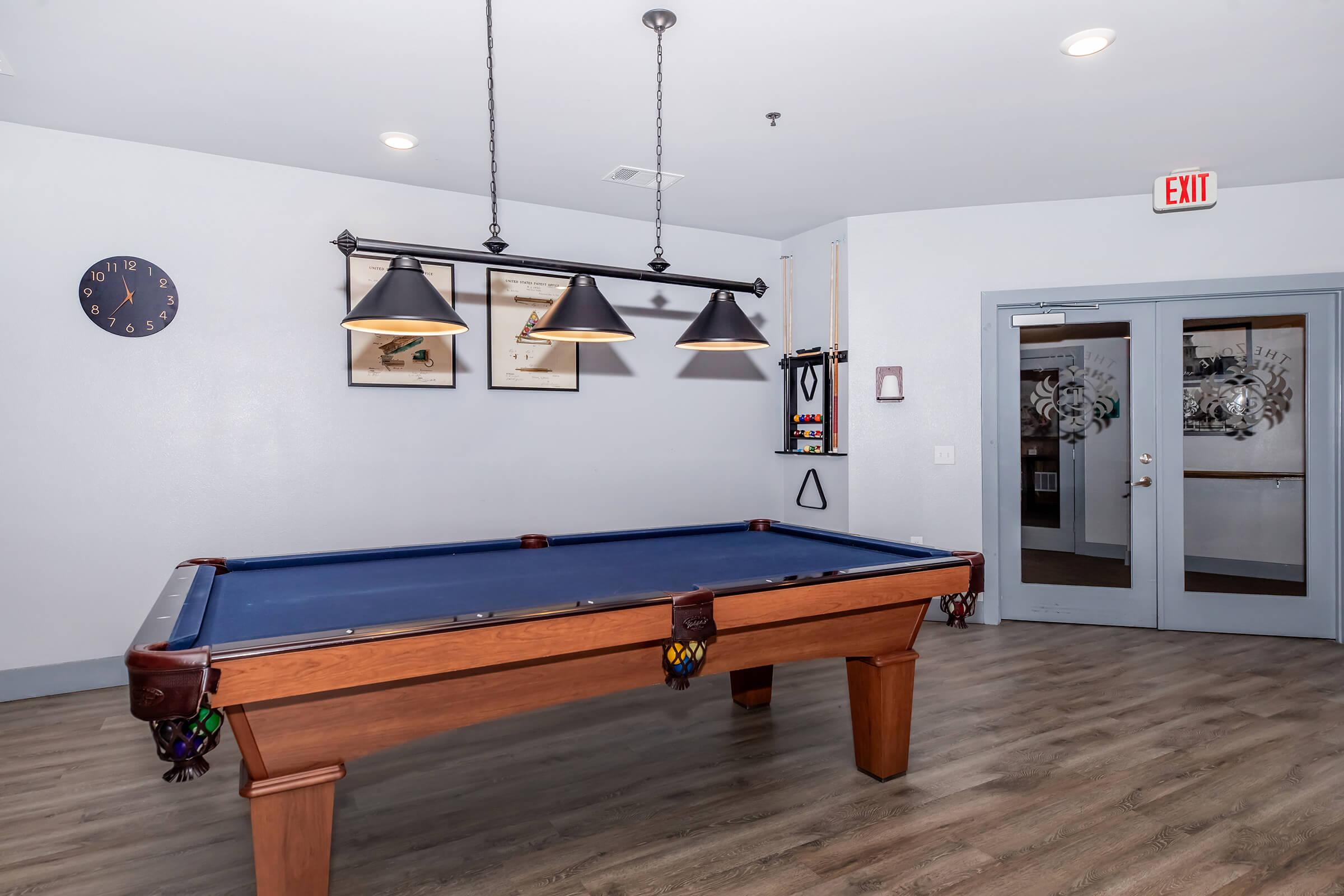 A well-lit recreational room featuring a blue felt pool table with decorative elements, including hanging pendant lights and a wall clock. The walls are adorned with framed artwork, and there are double doors leading to another area, creating a welcoming atmosphere for leisure activities.