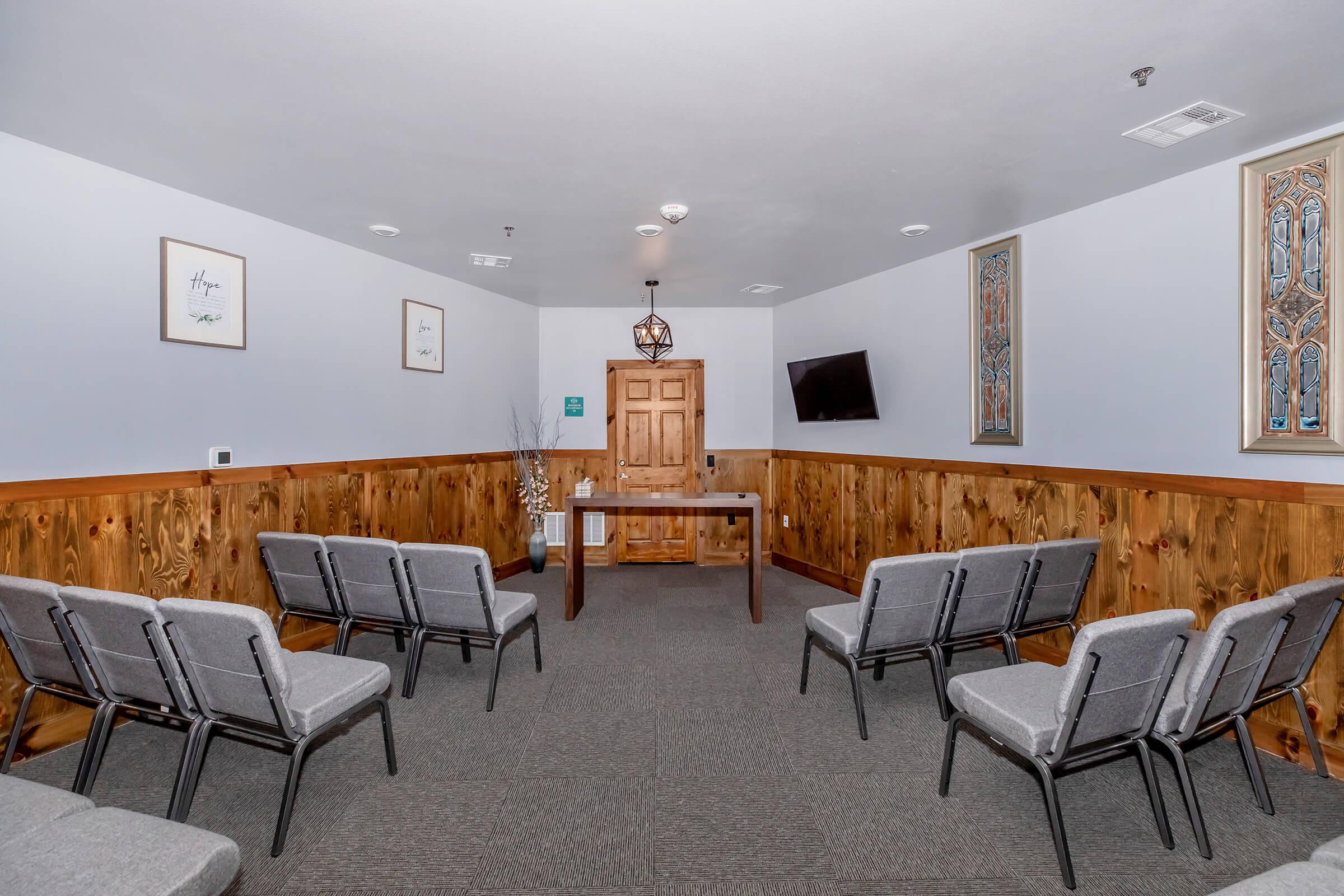 A simple room with gray chairs arranged in rows facing a wooden table. The walls are adorned with framed artwork and a decorative window. A TV is mounted on one wall, and there are wooden accents along the lower part of the walls, creating a warm and inviting atmosphere.
