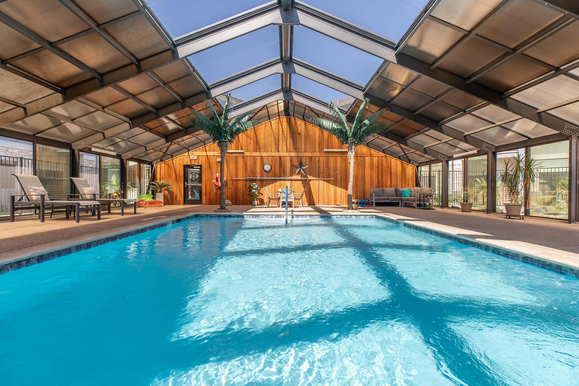 Indoor swimming pool area with a clear roof, surrounded by palm trees and lounge chairs. The pool water is calm and inviting, reflecting sunlight. A wooden wall provides a warm backdrop, while poolside amenities are visible in the background, creating a relaxing atmosphere.