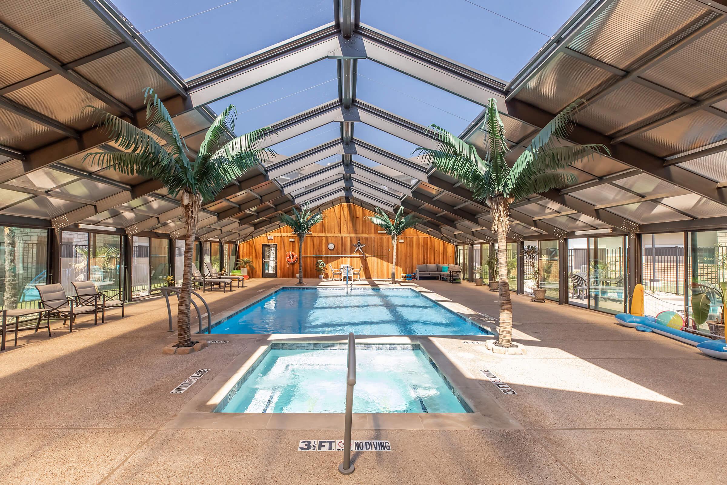Indoor swimming area featuring a large pool and a hot tub, surrounded by palm trees. The space is enclosed with glass panels, providing natural light. Seating areas are visible along the sides, and a wooden wall adds warmth to the contemporary design.