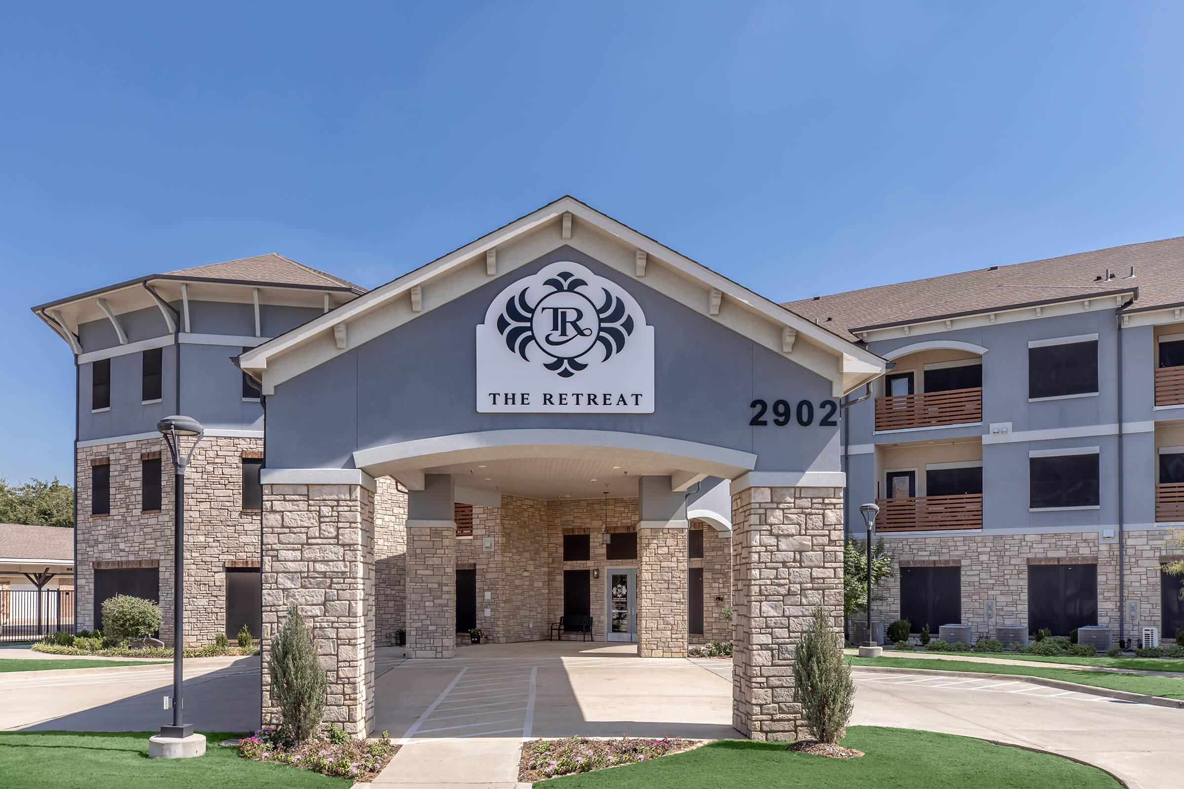 Exterior view of a building featuring a prominent entrance labeled "The Retreat." The structure has a modern design with a stone facade and decorative elements. Clear blue skies and landscaped greenery surround the entrance, providing a welcoming atmosphere.