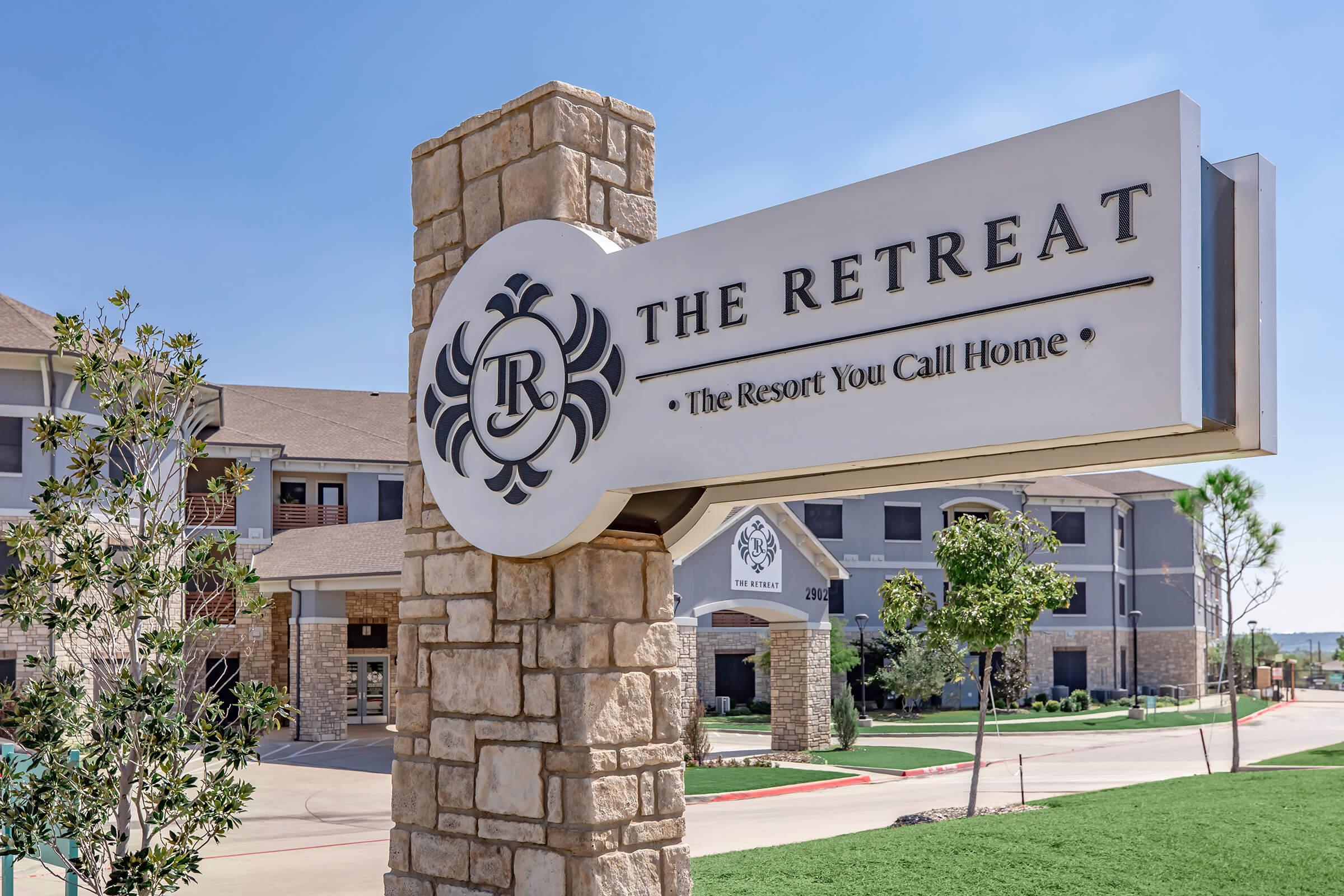 Sign for "The Retreat" with the tagline "The Resort You Call Home" in front of a modern building with a stone structure and landscaped surroundings. Clear blue sky adds to the inviting atmosphere of the resort.