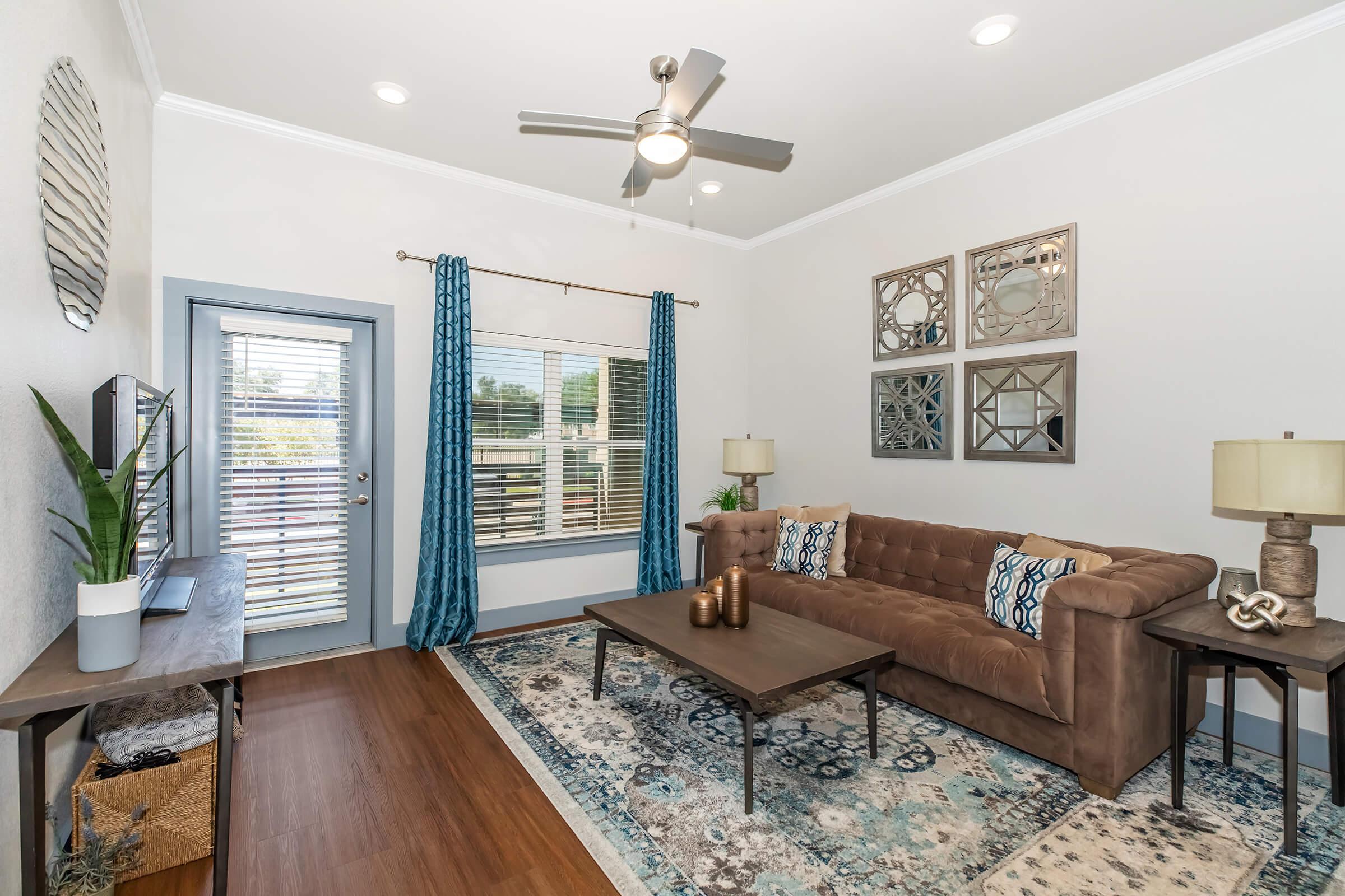 A cozy living room featuring a brown tufted sofa with decorative pillows, a wooden coffee table with decorative items, and a stylish blue area rug. Large windows with blinds let in natural light, complemented by light blue curtains. Wall decor includes framed mirrors and a round wall piece, creating a welcoming atmosphere.