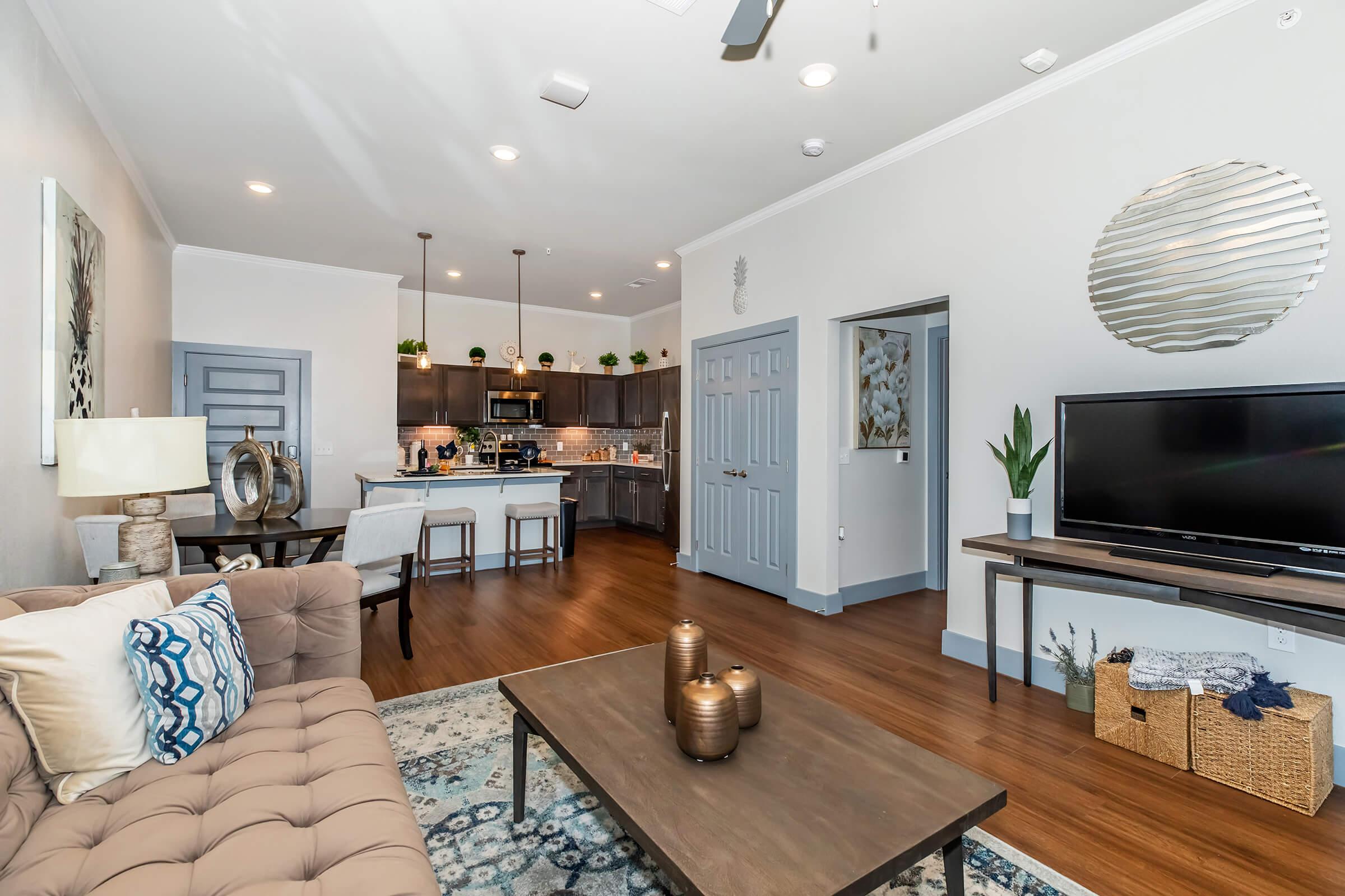 A modern, stylish living room with a beige sofa, a wooden coffee table, and decorative items. The open-concept space leads to a kitchen with dark cabinetry and a breakfast bar. A wall-mounted TV and a decorative round mirror are visible, along with potted plants and cozy decor, creating an inviting atmosphere.