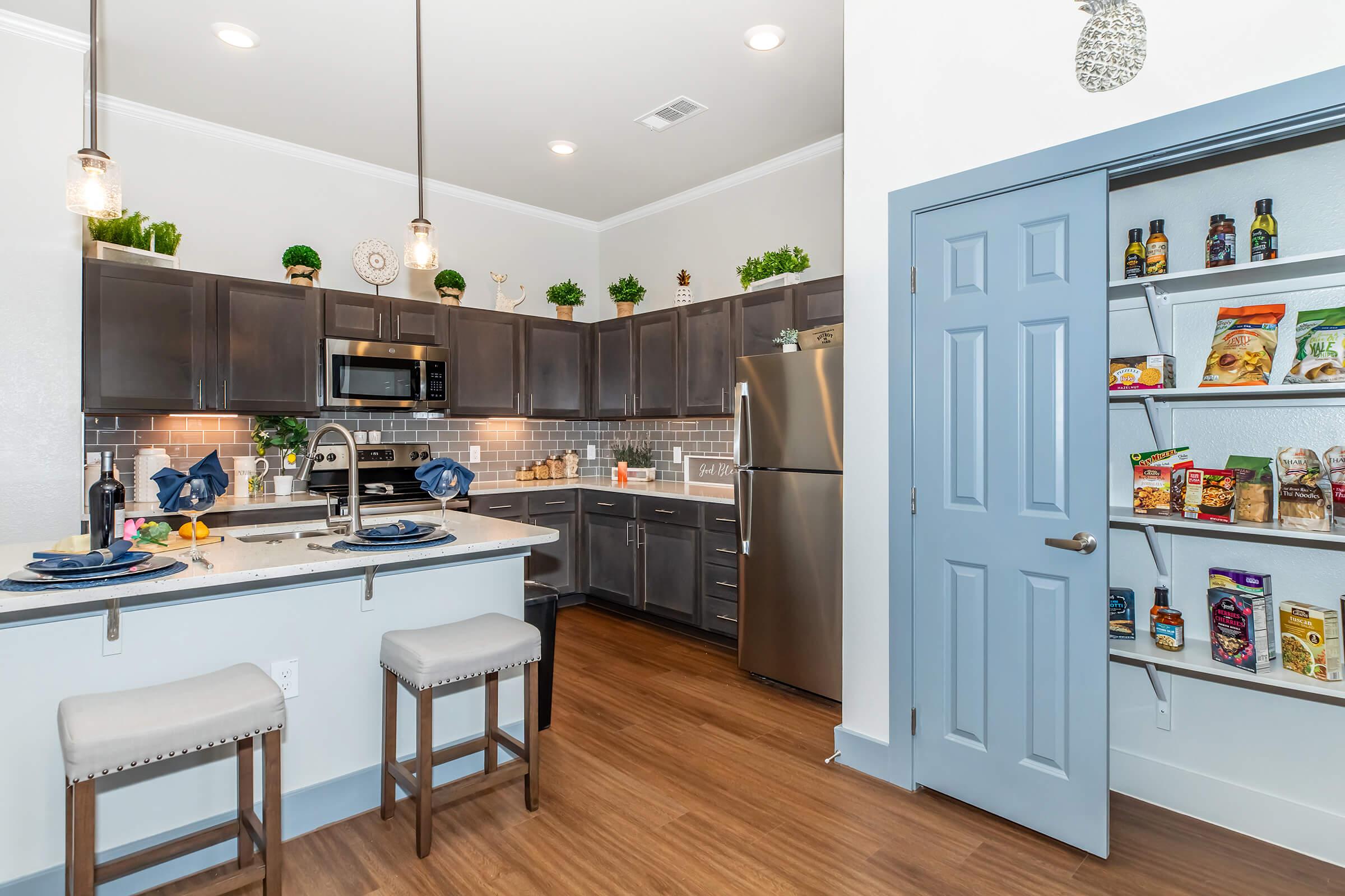 Modern kitchen featuring dark wood cabinetry, stainless steel appliances, and a gray tile backsplash. A central island with bar stools is set for dining. A pantry door is open, showcasing organized shelves with food items. Soft lighting highlights the space, creating a warm and inviting atmosphere.