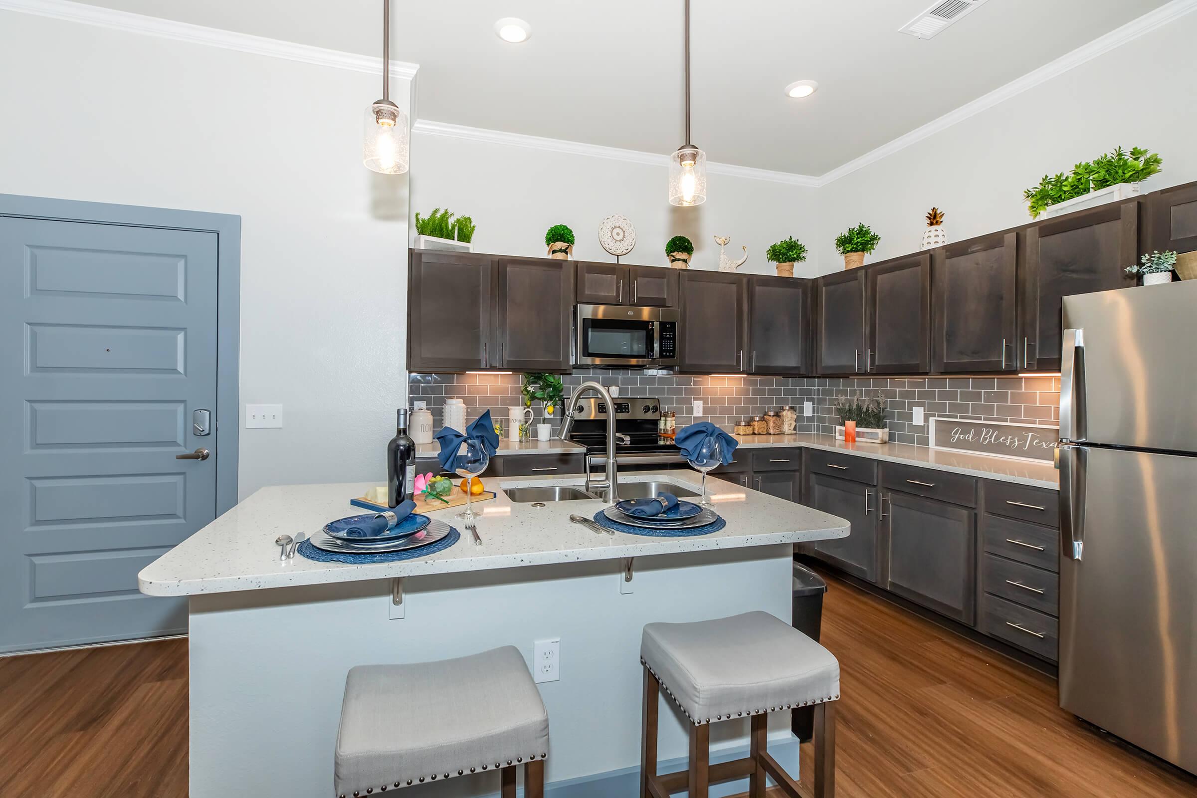 Modern kitchen featuring dark wood cabinetry, stainless steel appliances, and a central island with bar seating. The space is well-lit with pendant lights and decorated with potted plants. A stylish table setting is visible on the countertop, complemented by warm wood flooring and an inviting aesthetic.