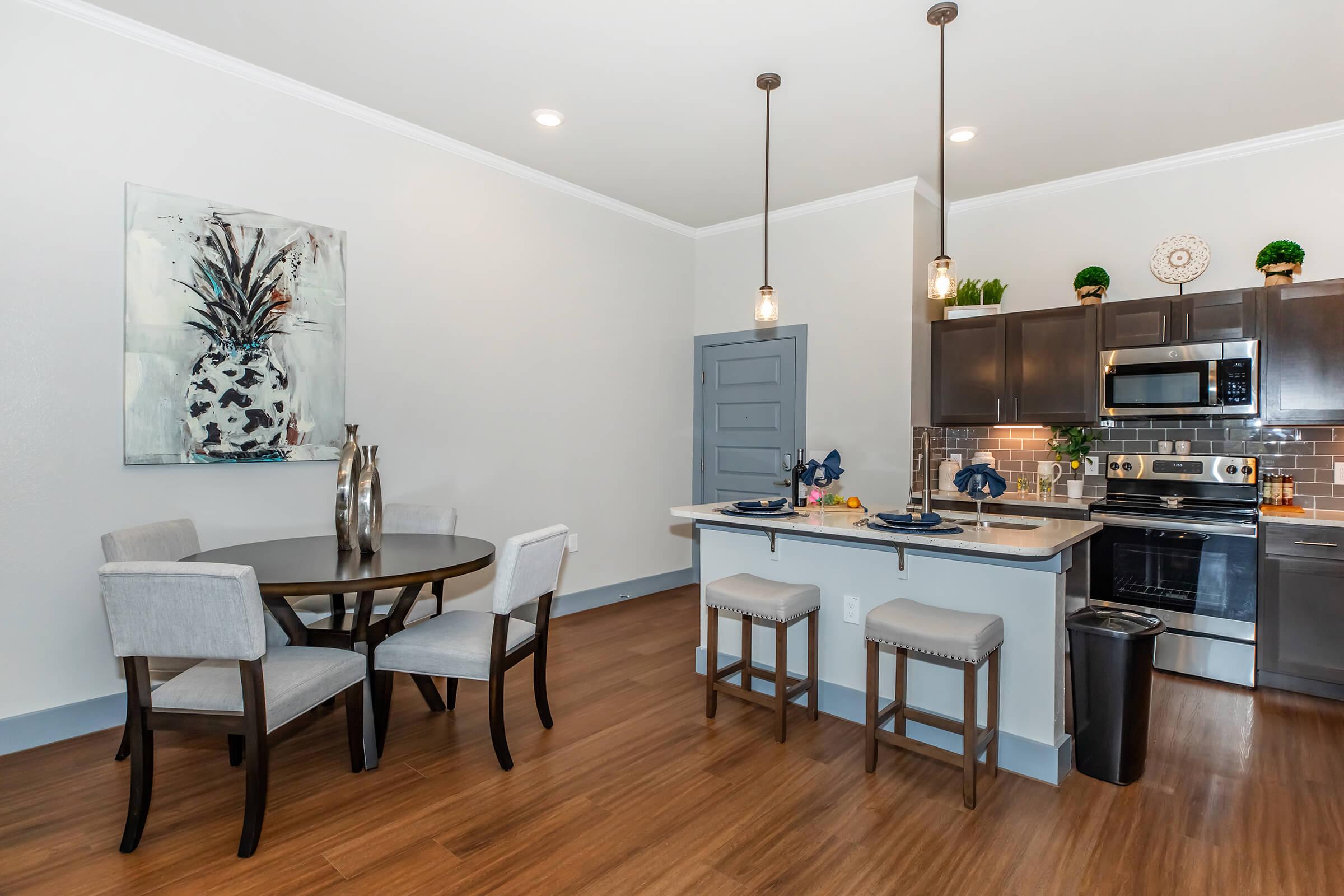 Modern kitchen and dining area featuring a round table with four upholstered chairs, a stylish vase on the table, dark cabinetry, stainless steel appliances, and pendant lighting. The walls are painted light gray with a decorative wall art piece of a plant in a vase. Wood flooring completes the contemporary look.