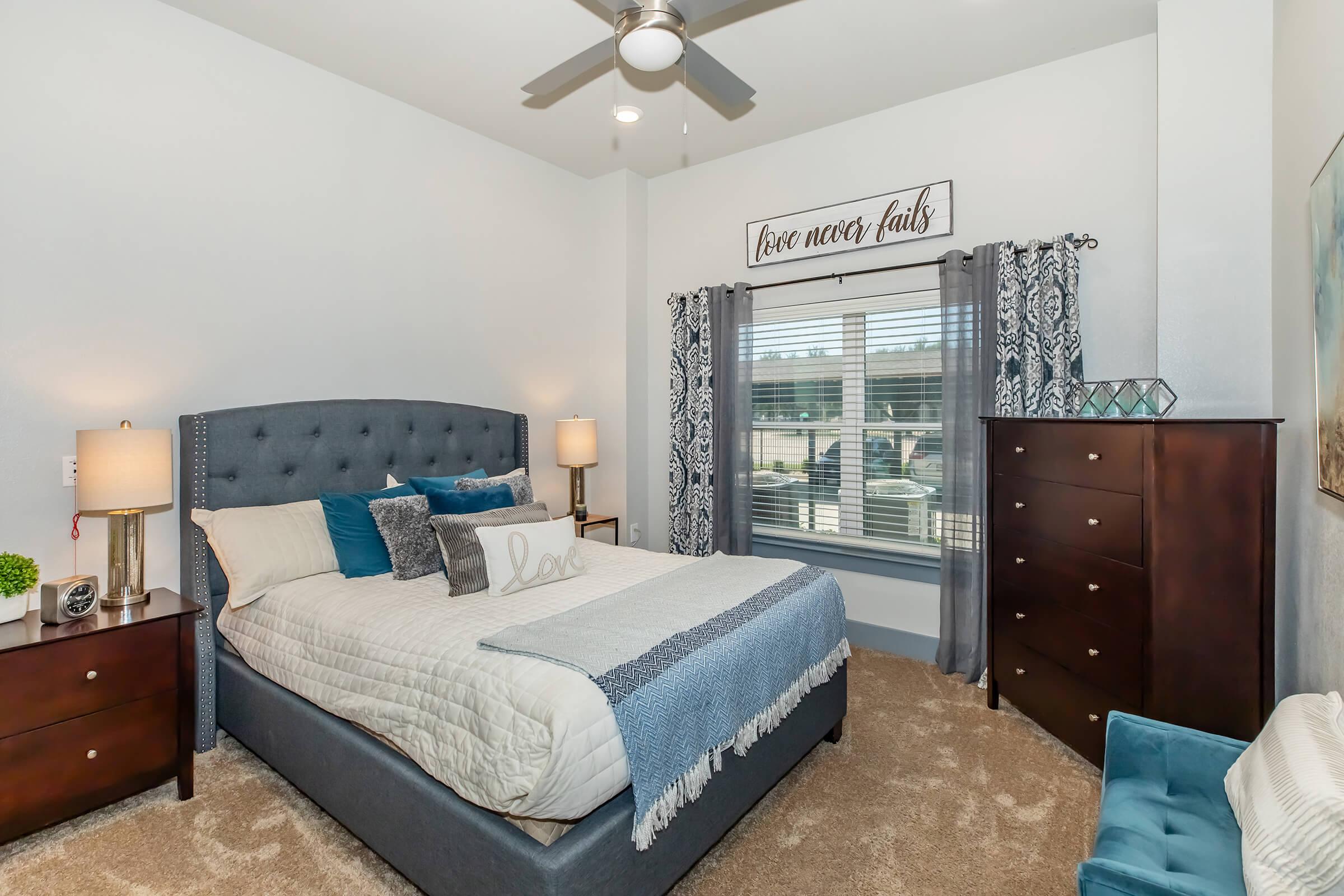 Cozy bedroom featuring a large bed with decorative pillows, a stylish headboard, nightstands with lamps on either side, a wooden dresser, a comfortable chair, and elegant window curtains. The room is well-lit with natural light and has a calming color palette.