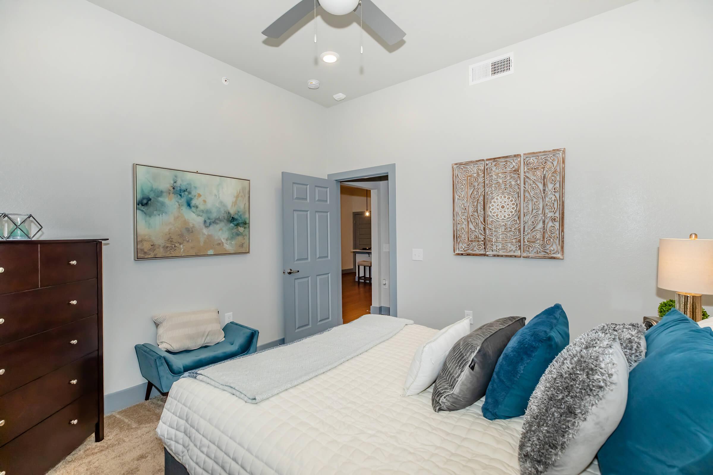 A cozy bedroom featuring a queen-sized bed with decorative pillows, a dark wooden dresser, and an armchair in the corner. The walls are painted light gray, and there's a piece of abstract art and a decorative wall hanging. A ceiling fan and natural light enhance the ambiance.