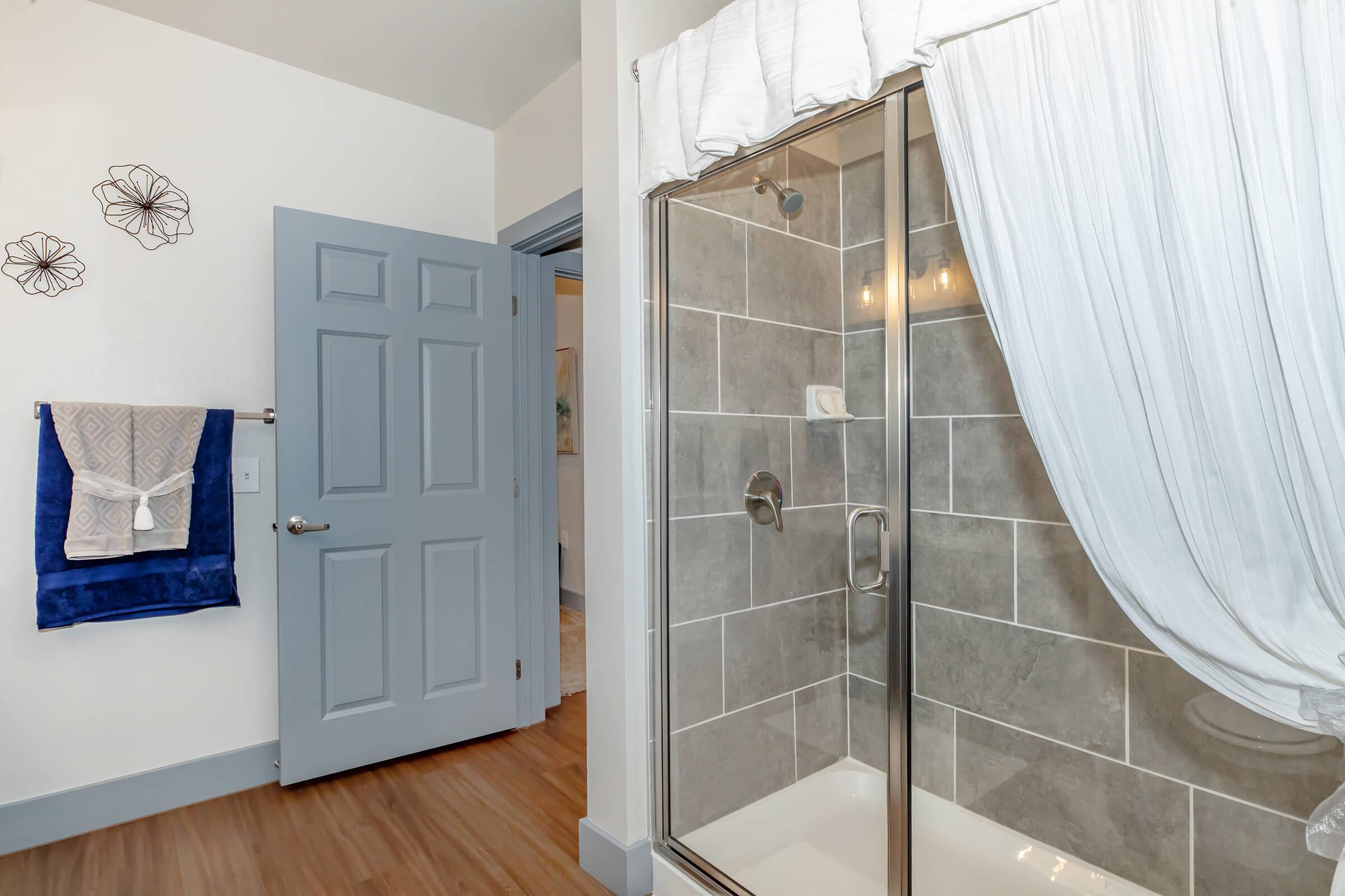 A modern bathroom featuring a glass shower enclosure, a decorative white curtain, and a blue towel hanging on a hook. The walls are adorned with flower patterns, and a light gray door leads to another room. The flooring is a warm wood tone, creating a welcoming atmosphere.