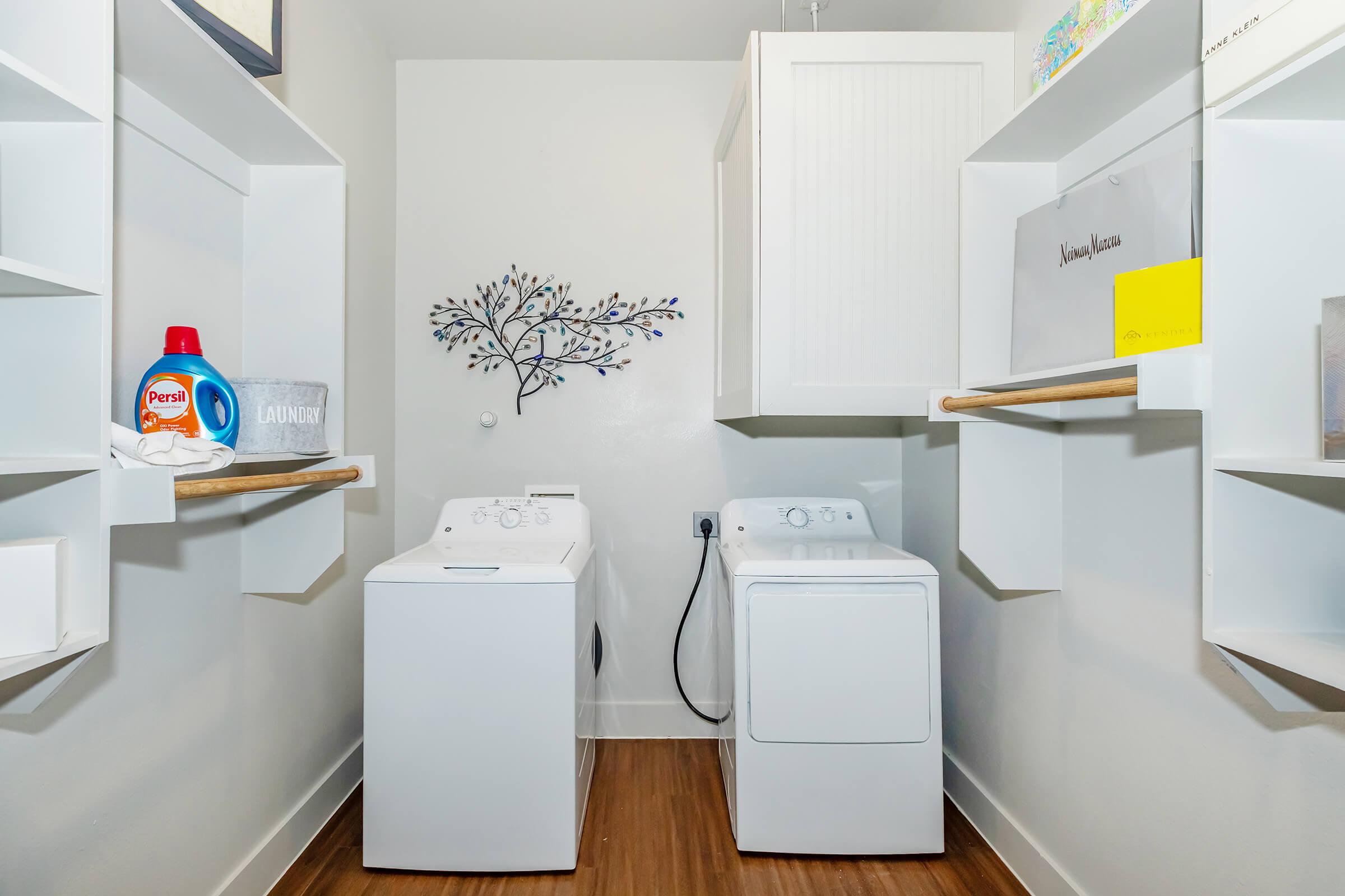 A clean and modern laundry room featuring a white washing machine and dryer side by side, with wooden shelves above them. The shelves hold decorative items and storage boxes. A wall decoration resembling a tree can be seen, adding a stylish touch to the space.