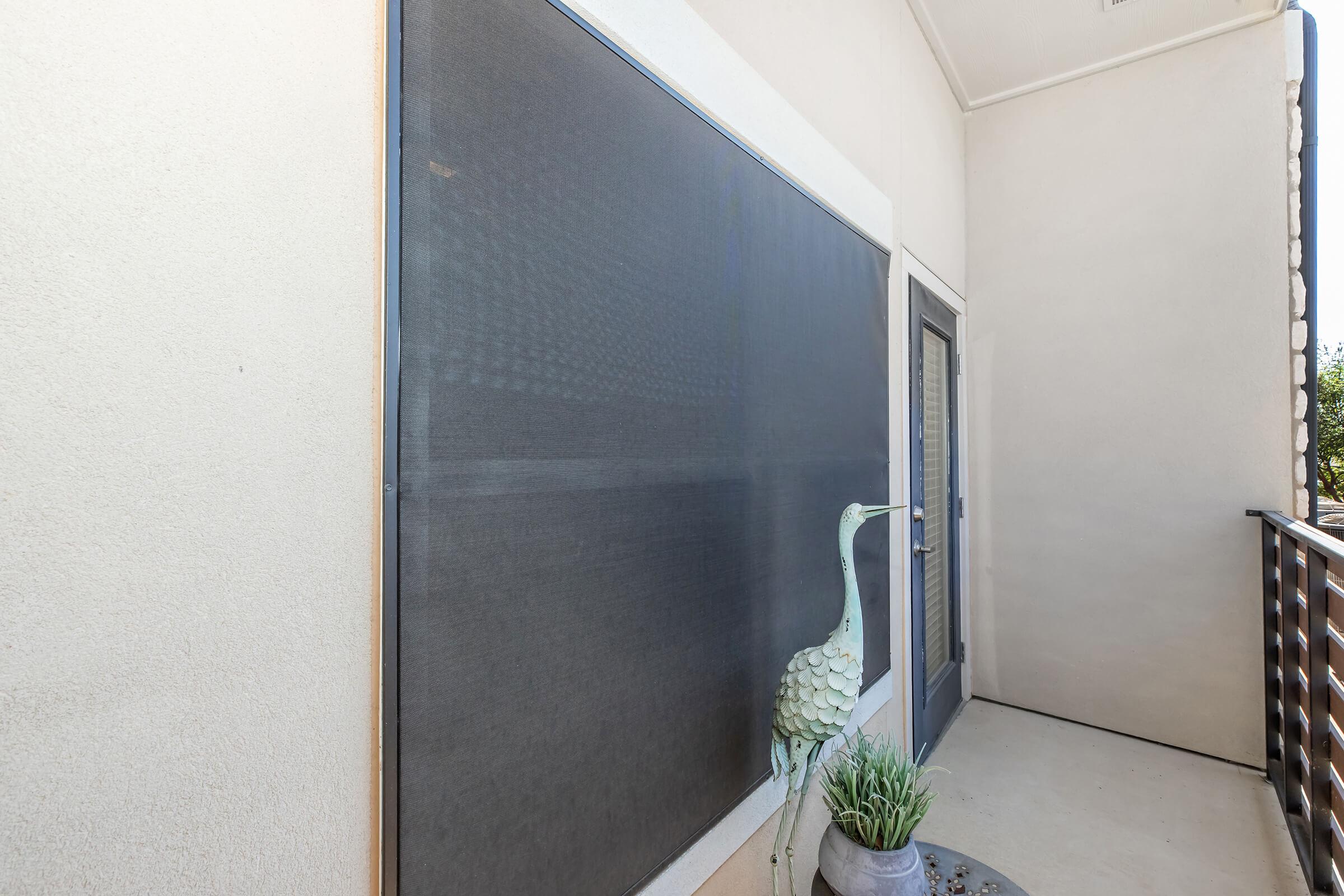 A balcony with a large screen on one wall, a door leading inside, and a decorative metal crane statue beside a potted plant. The balcony features a clean, modern design with neutral-colored walls and a simple railing.