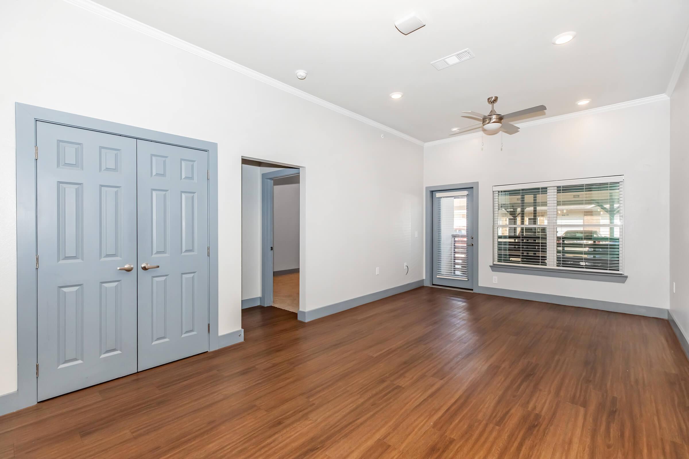 Bright and spacious living room featuring light wood flooring, a ceiling fan, and large windows with blinds. A blue closet door and another doorway lead to additional rooms. The walls are painted in a light neutral color, enhancing the open and airy feel of the space.