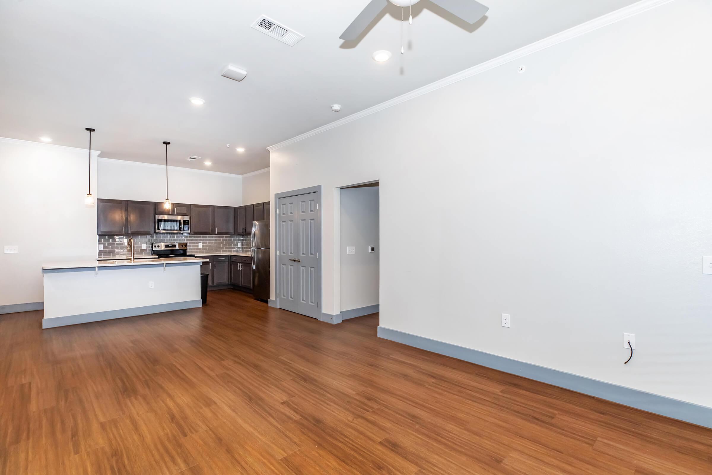 Modern open-concept living space featuring a kitchen with dark cabinetry and stainless steel appliances. Light-colored walls and hardwood flooring create a bright atmosphere. Ceiling fan and recessed lighting add comfort and style. A doorway leads to another room, enhancing the layout.