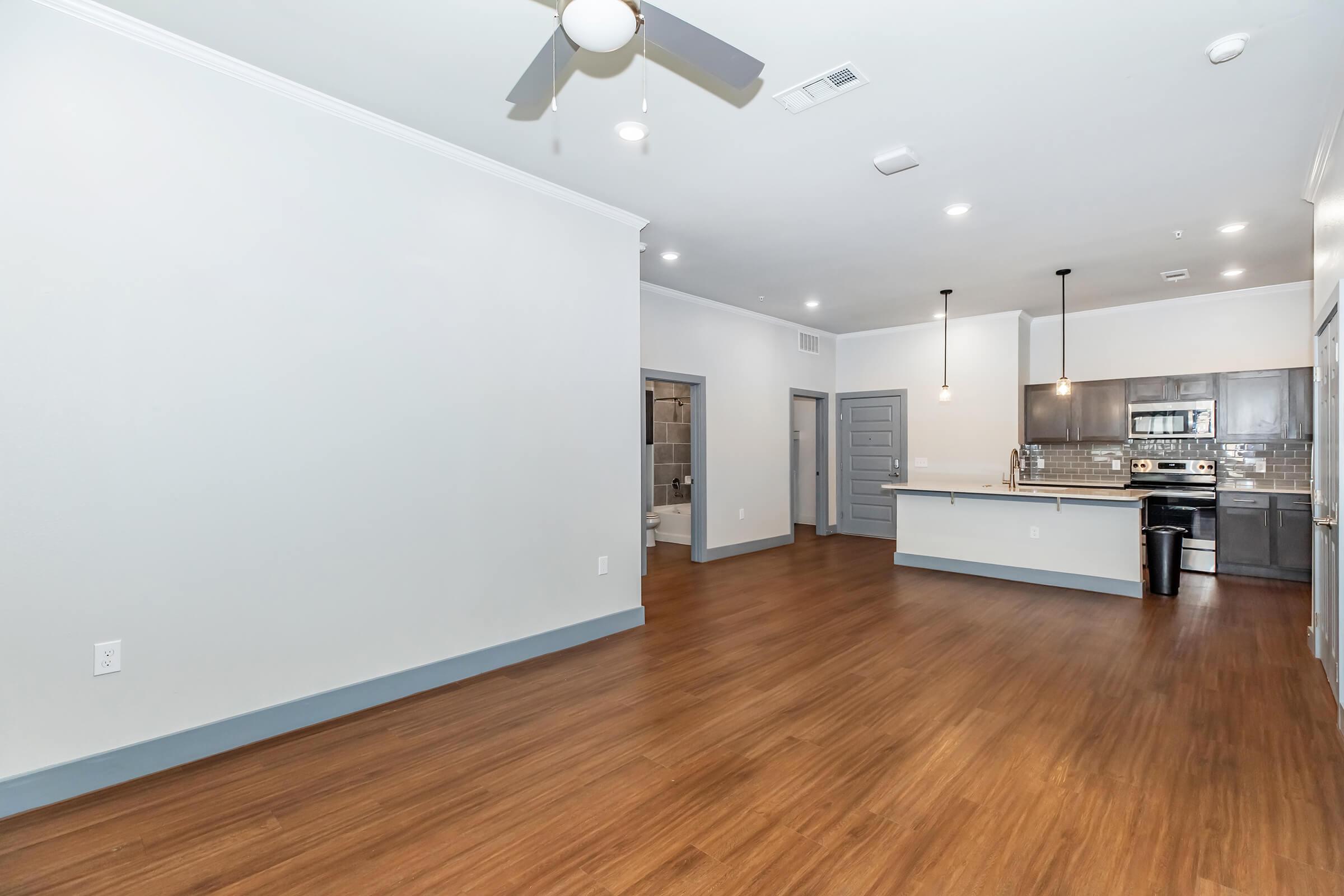 Spacious modern living room with hardwood floors, ceiling fan, and open kitchen featuring stainless steel appliances. Neutral walls and lighting create a bright atmosphere. Additional doorways lead to other areas, enhancing the living space's flow and accessibility.