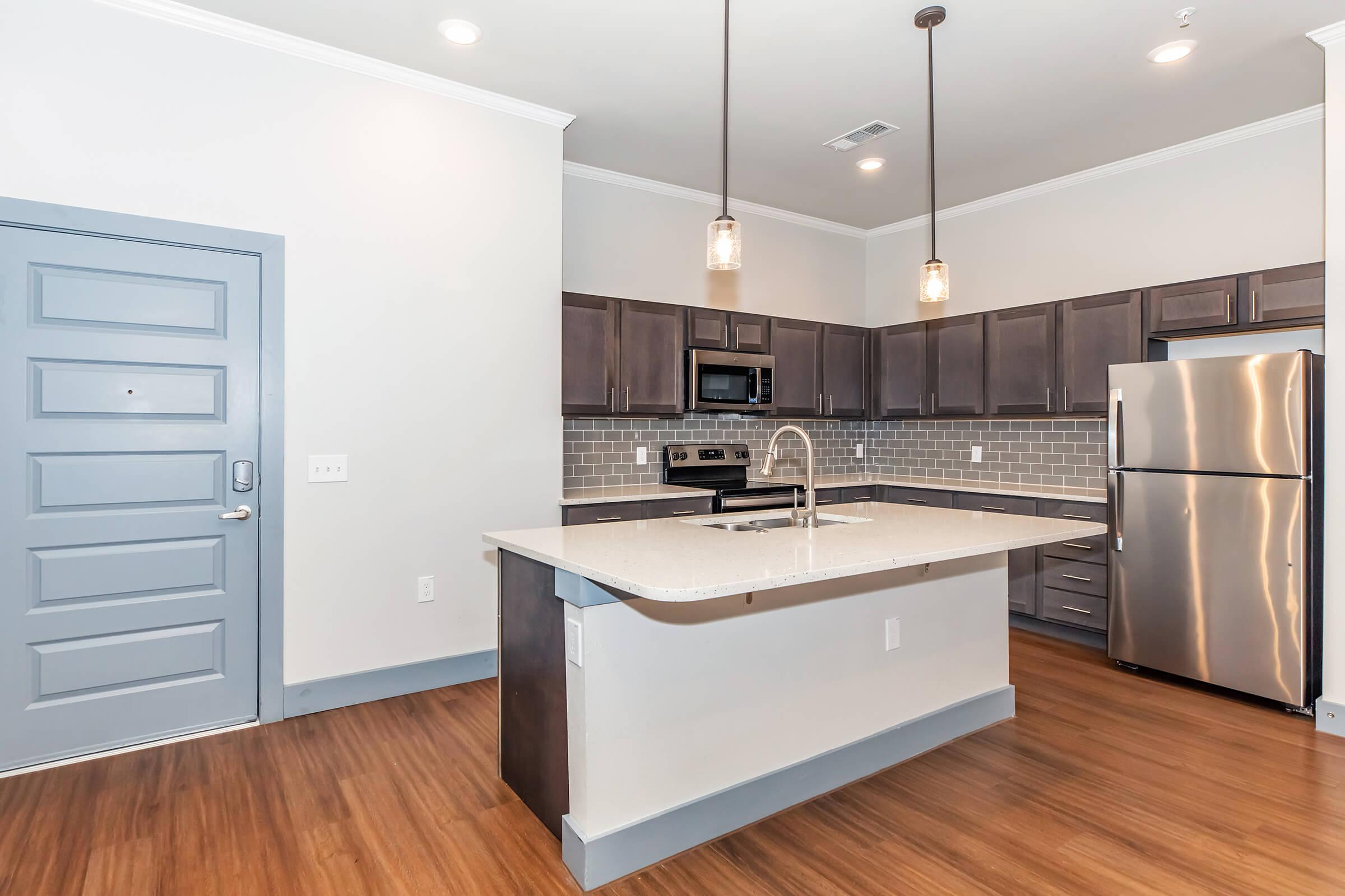 Modern kitchen in an apartment featuring dark wood cabinetry, stainless steel appliances including a refrigerator and oven, a large island with a white countertop, and stylish pendant lighting. The flooring is a warm wood laminate and the walls are painted in a soft gray.