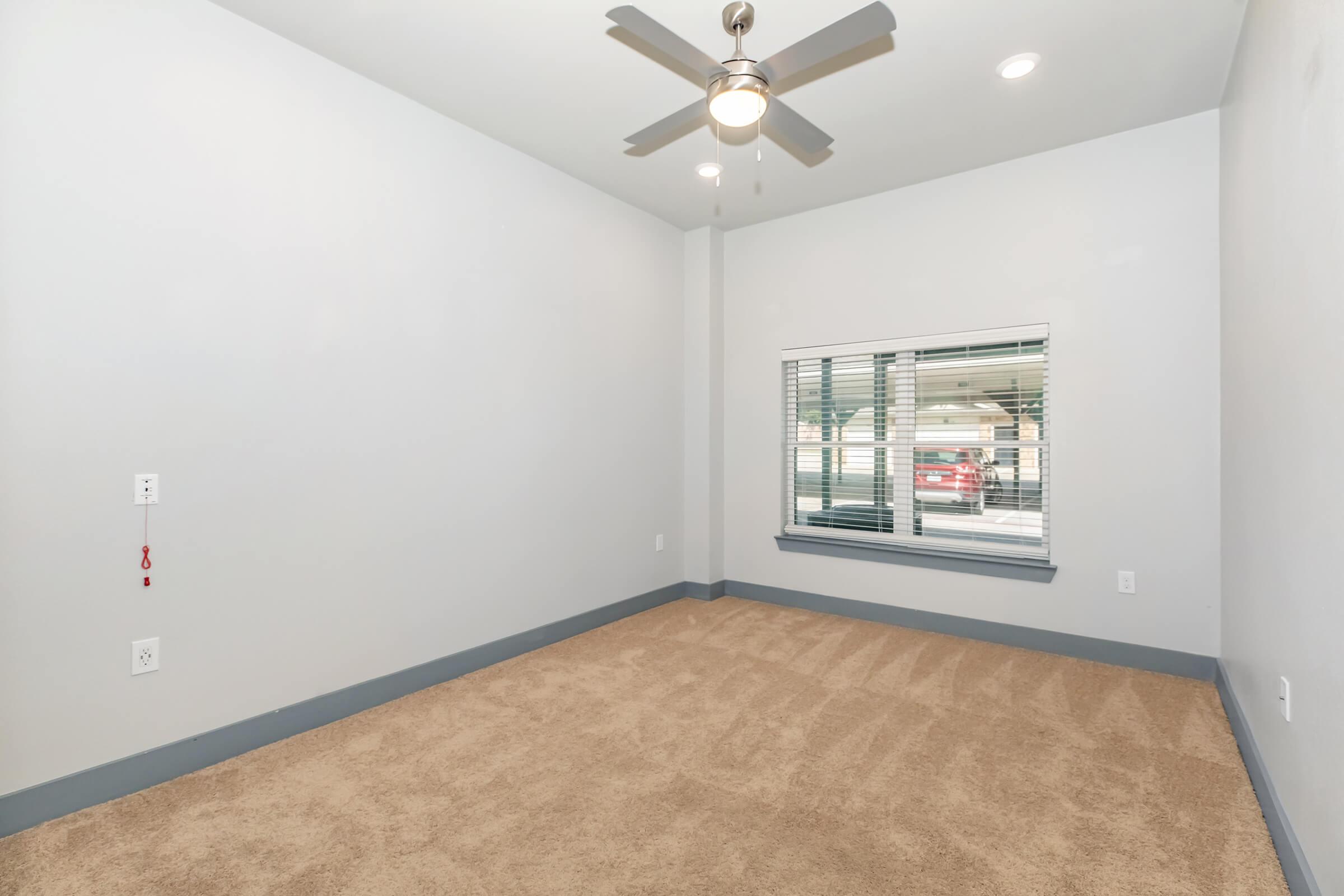 Empty room with beige carpet, light gray walls, and a ceiling fan. A window with white blinds overlooks an outdoor area, providing natural light. The room features a minimalistic design with no furniture or decorations.