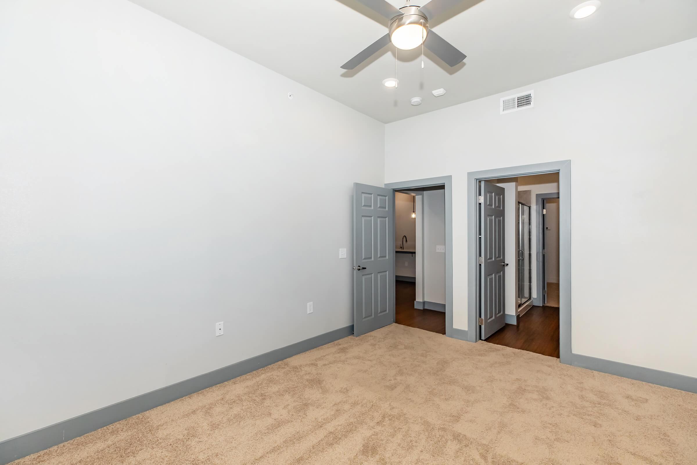 A spacious room with light gray walls and brown carpet flooring. It features a ceiling fan and two open doorways leading to other areas. The lighting is bright and the overall decor is modern and minimalistic.