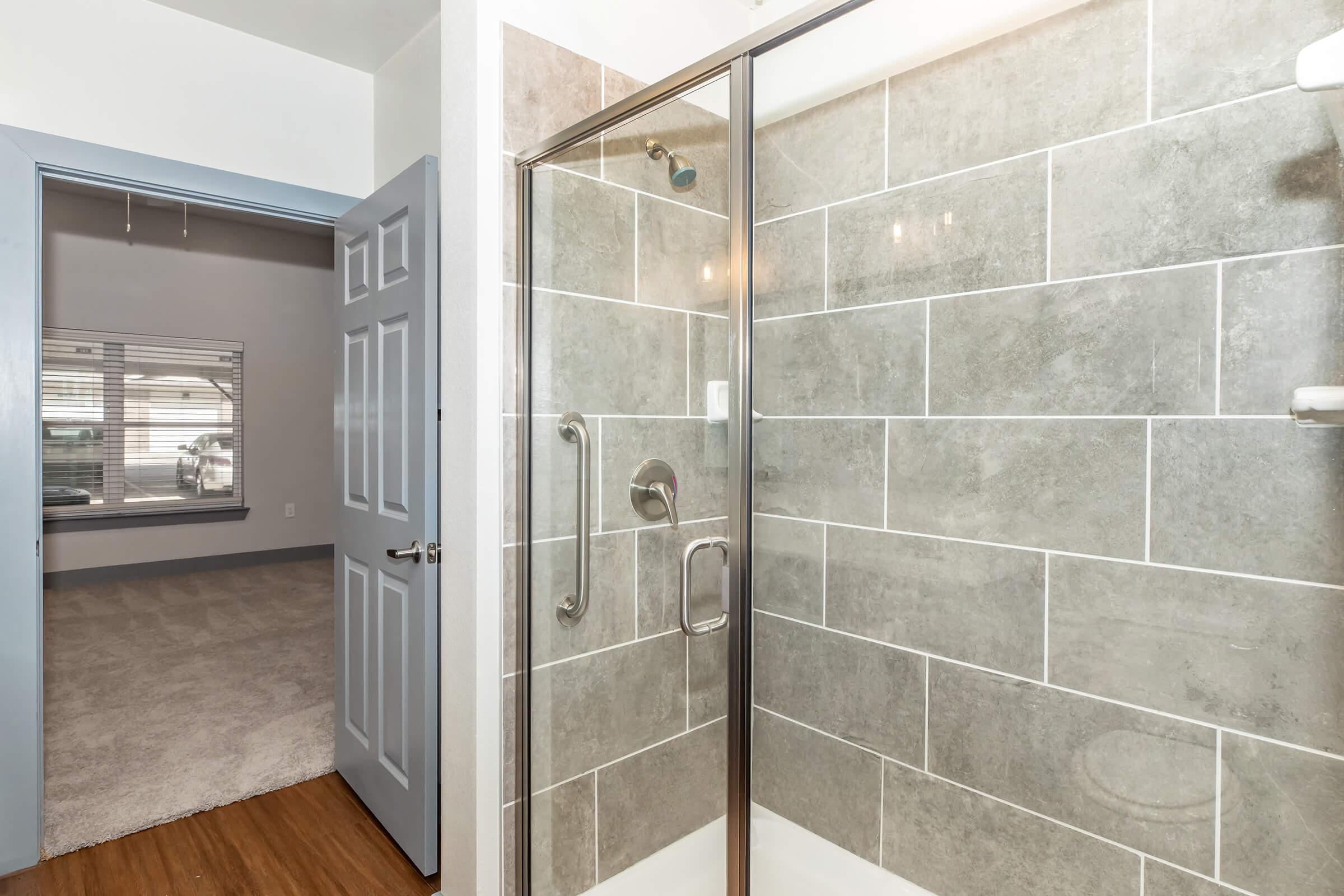 Modern bathroom featuring a glass shower enclosure with gray tiled walls, adjacent to a room with light gray carpet and a window. A blue door leads to the room, enhancing the clean and contemporary aesthetic.