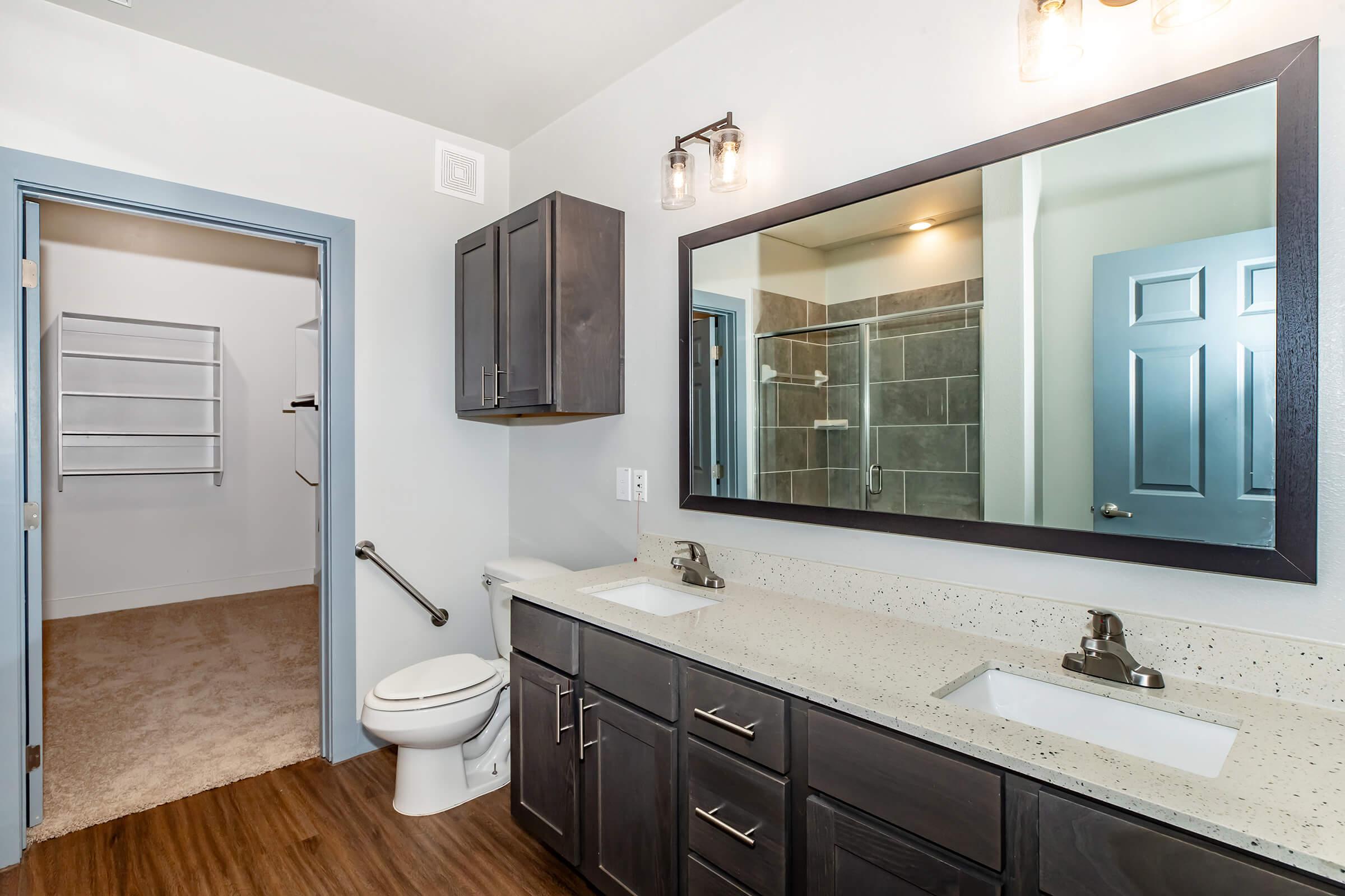 A modern bathroom featuring a double vanity with two sinks, dark cabinetry, and a large mirror above. There is a toilet on the left side, and a walk-in shower with gray tiles is visible in the background. A doorway leads to a separate room with shelves, and the flooring is a wood-like laminate.