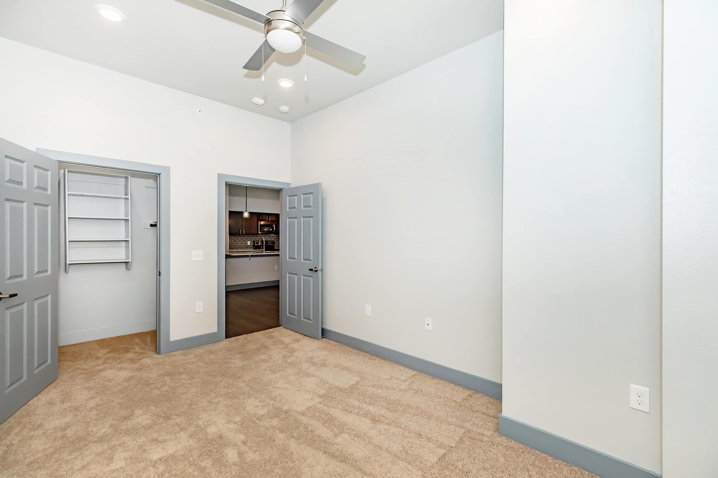 A bright, vacant room featuring beige carpet flooring, light gray walls, and a ceiling fan. There are two open doors leading to closets and a glimpse of a kitchen area in the background. The room is well-lit, emphasizing its spaciousness and clean lines.