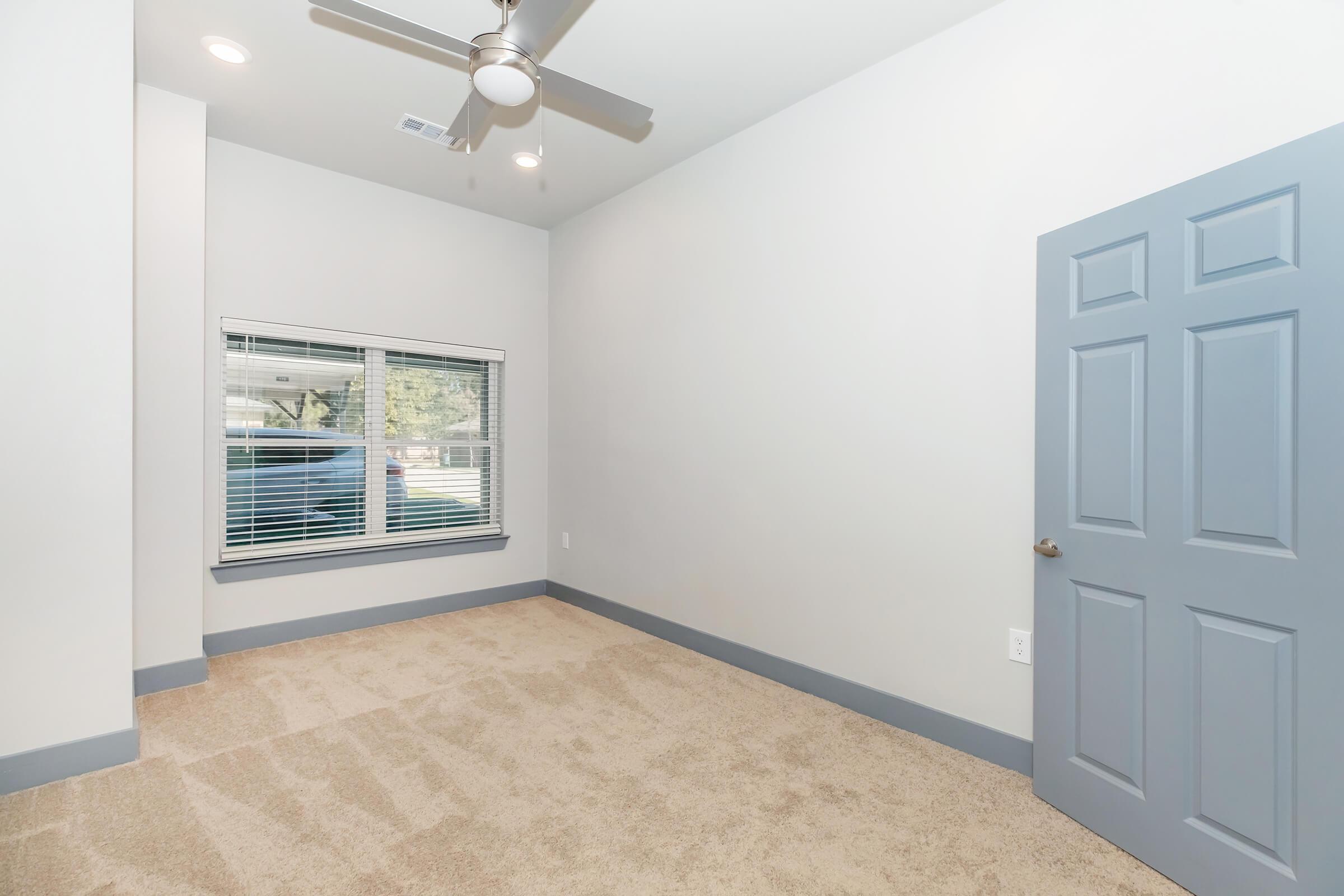 A brightly lit and spacious room with gray walls and light beige carpet. The room features a ceiling fan and a large window with blinds. A closed blue door is visible on the right side, enhancing the minimalistic design of the space.