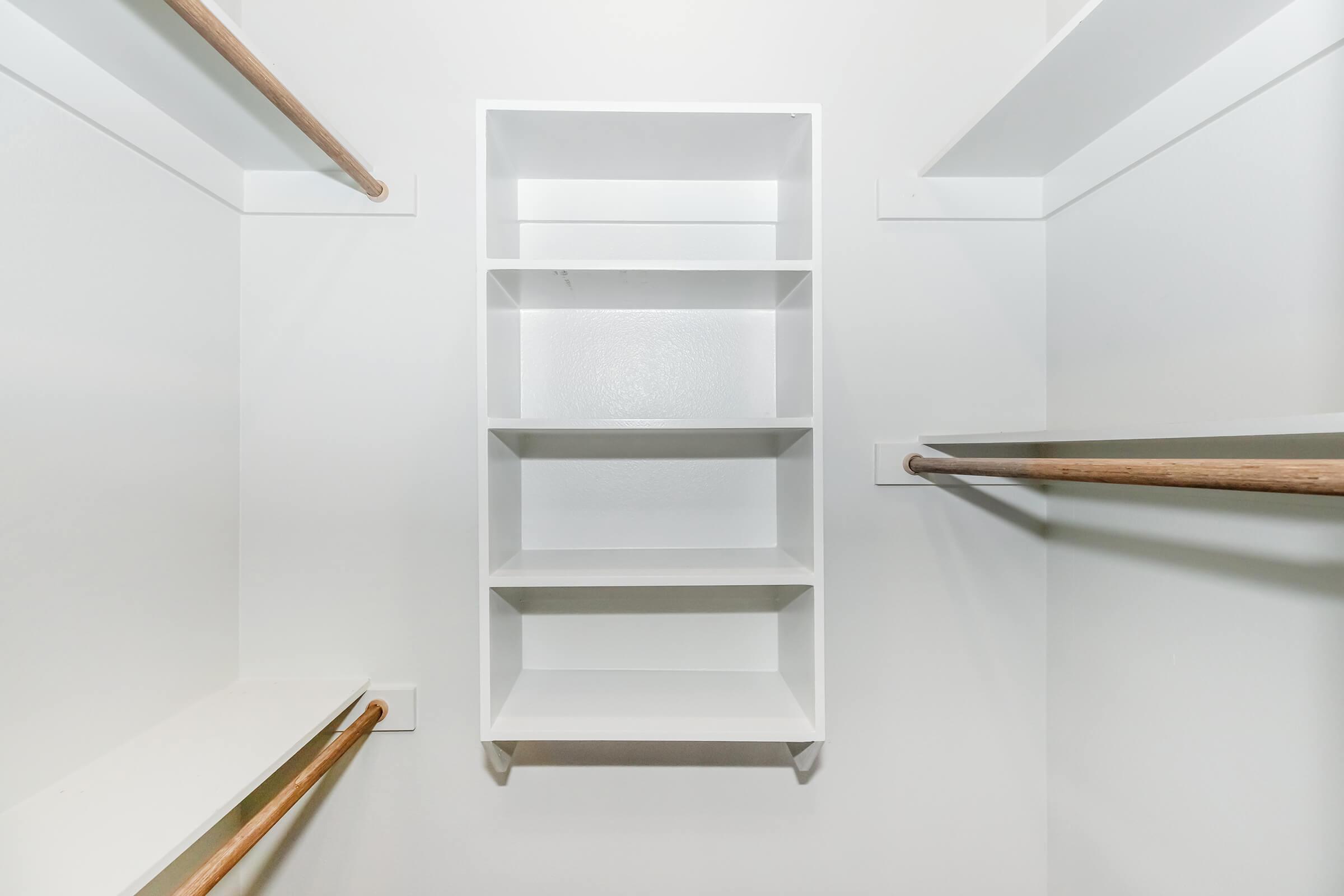 An empty white closet with a simple shelf unit in the center and two wooden rods on either side for hanging clothes. The walls and shelves are bright and uncluttered, creating a clean and minimalist appearance.