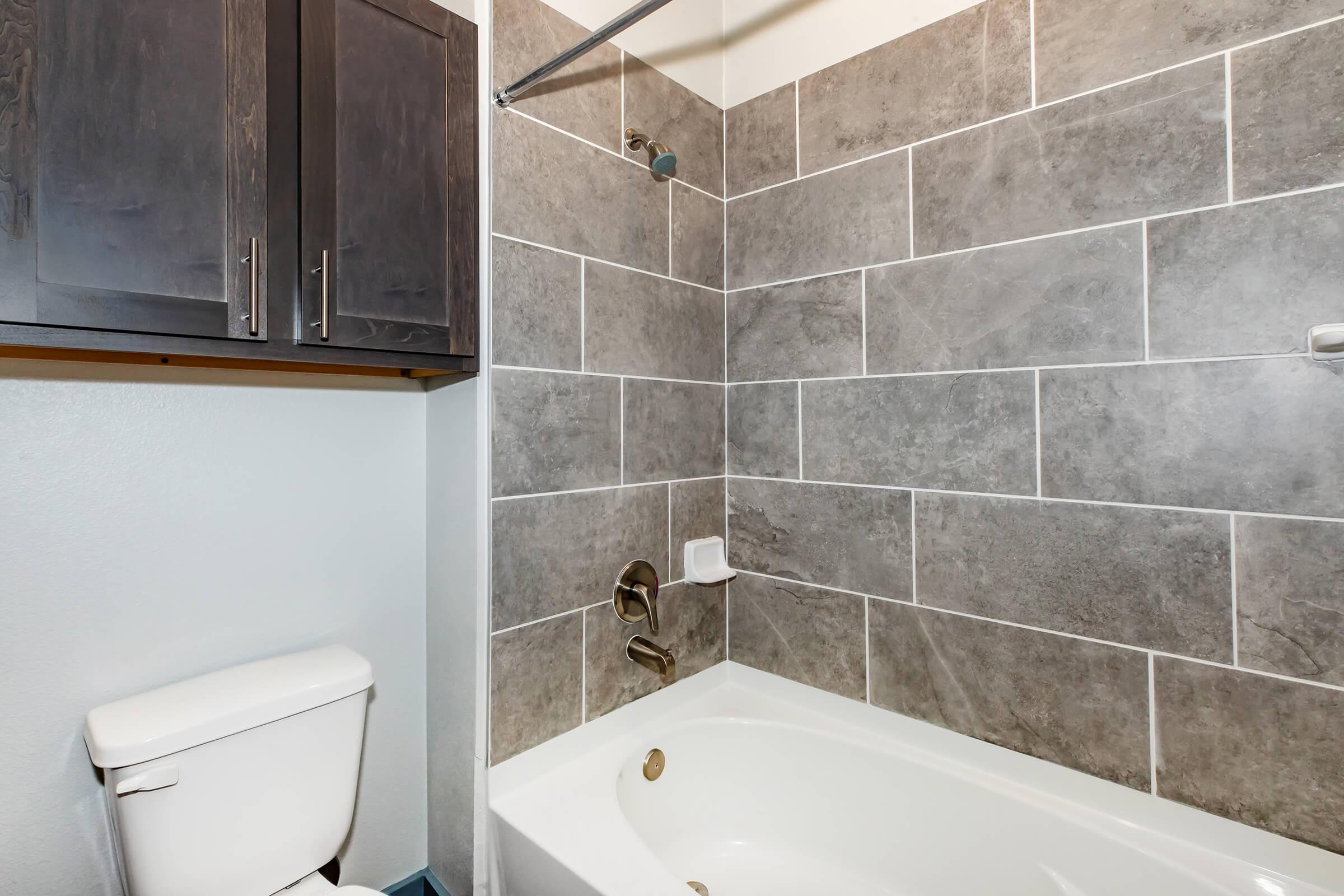 A modern bathroom featuring a white bathtub with a showerhead above, illuminated by natural light. The walls are tiled with large gray tiles, and there is a dark cabinet above the toilet for storage. The decor presents a clean and minimalist aesthetic.