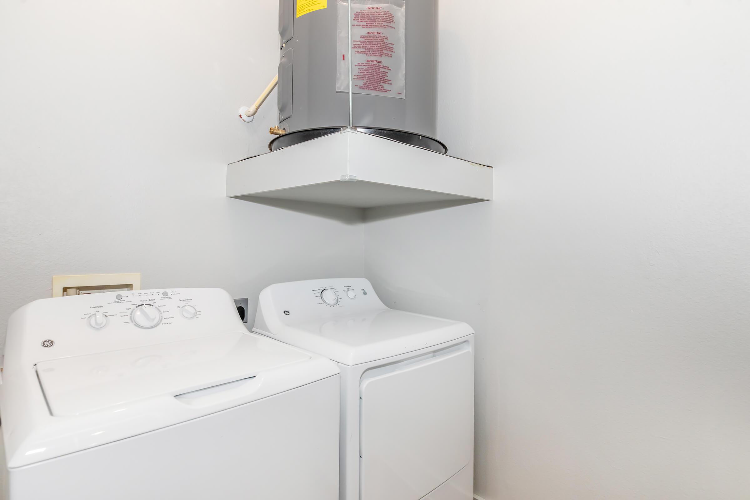 A clean and organized laundry room featuring a stacked washer and dryer in white, positioned on a shelf below a water heater. The walls are plain and light-colored, creating a bright and simple atmosphere.