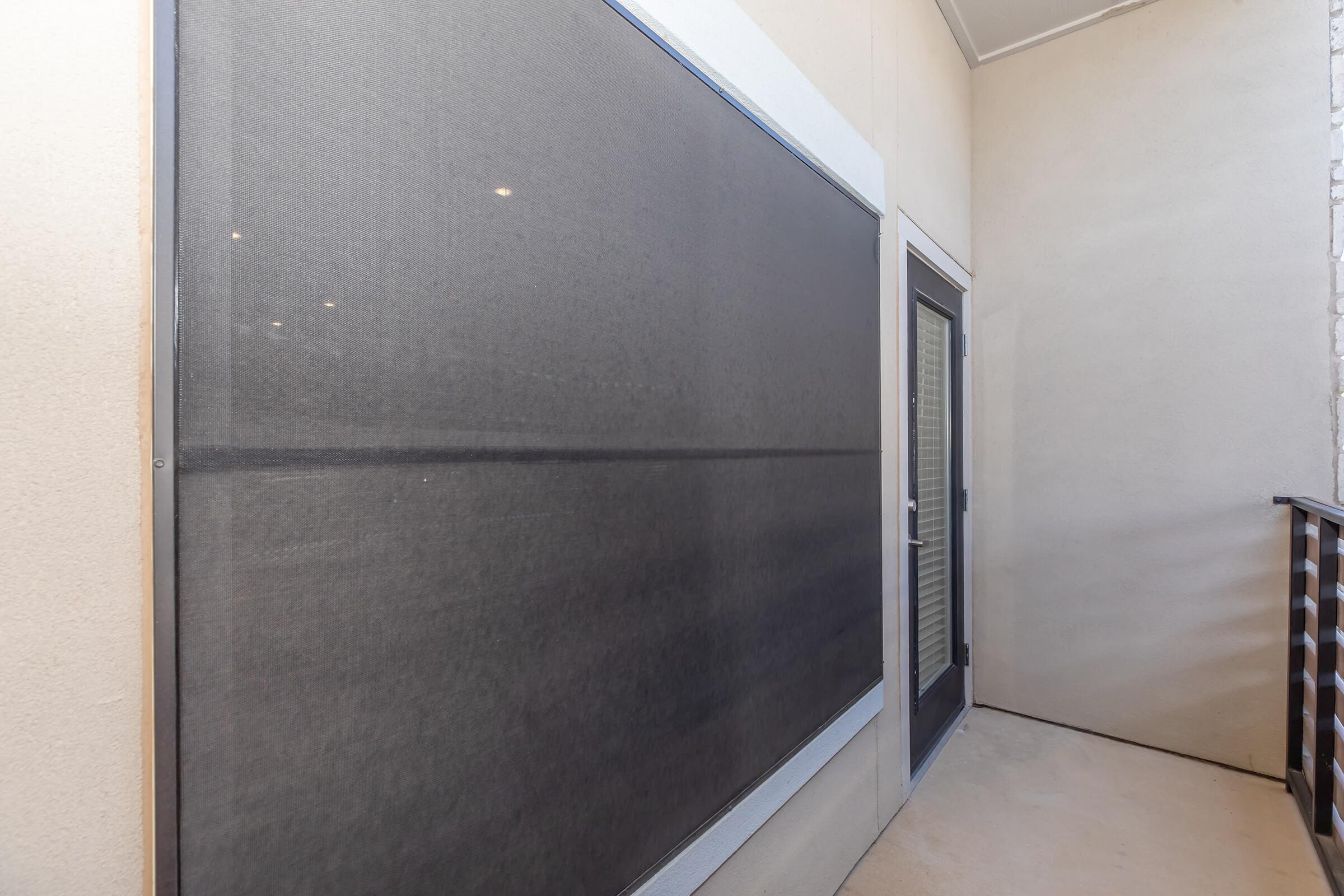 A balcony area featuring a large screen or mesh panel on one wall, with a door leading inside. The walls are beige and the floor is concrete, creating a simple, minimalistic outdoor space. The overall impression is one of a private balcony with limited visibility to the outside.