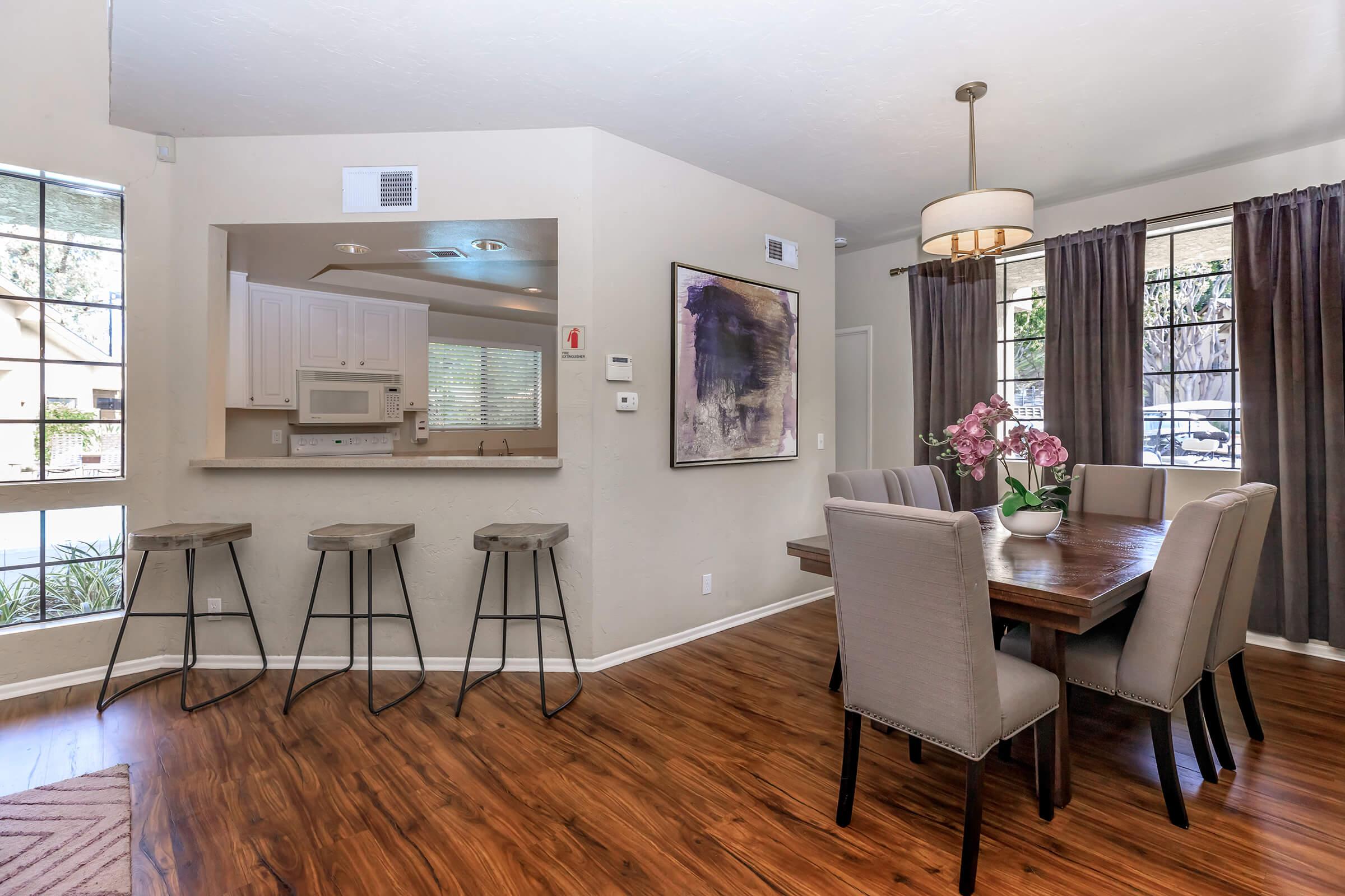 A modern dining area featuring a wooden table with six upholstered chairs, a decorative table centerpiece of purple flowers, and a stylish light fixture above. The adjacent kitchen bar has four high stools. Large windows allow natural light, complementing the warm wood flooring and neutral wall colors.