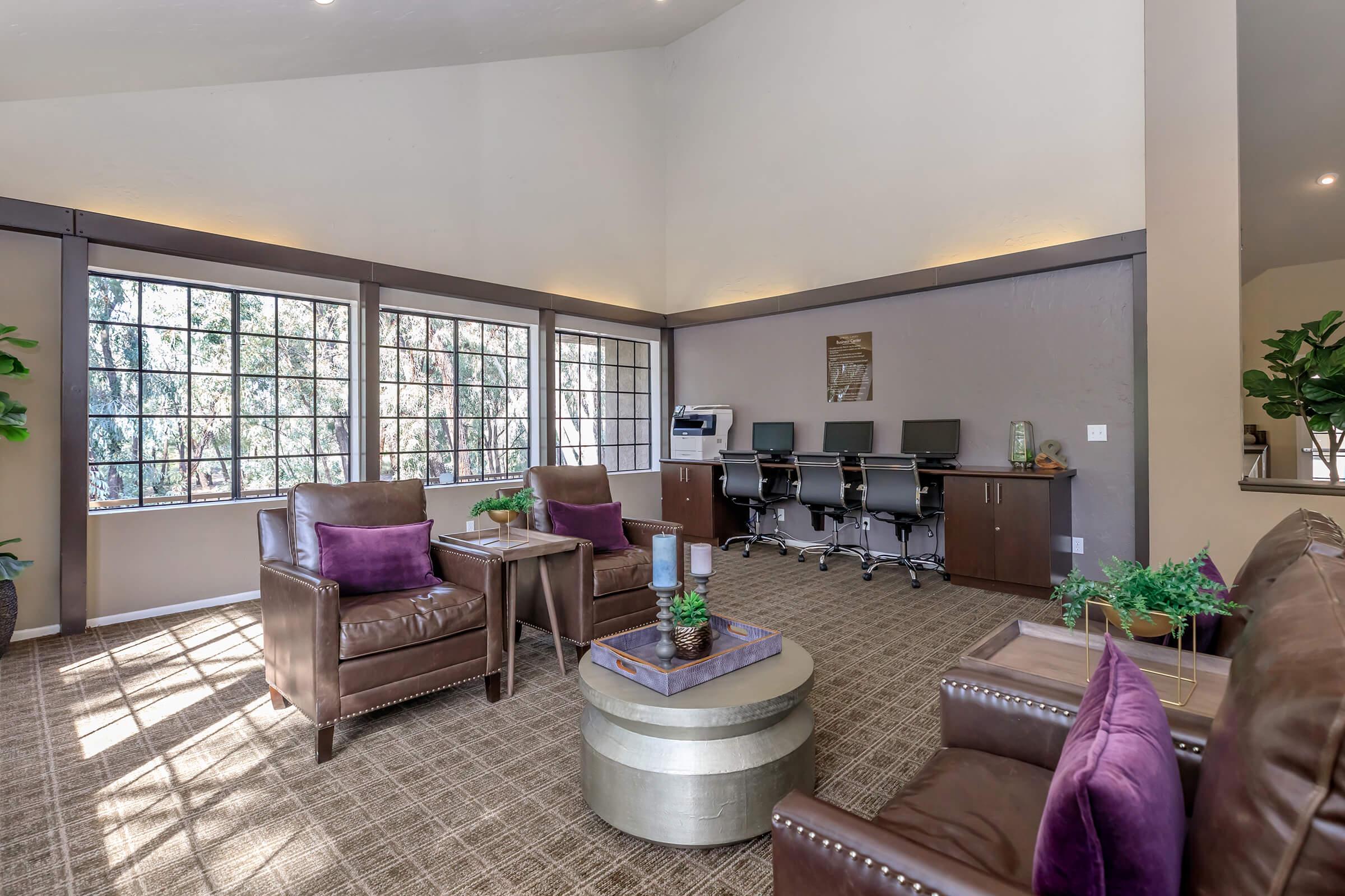 A cozy lounge area featuring two brown leather chairs with purple cushions, a round metallic table, and a desk with computers in the background. The space is well-lit by natural light coming through large windows, with greenery adding a touch of nature to the environment.