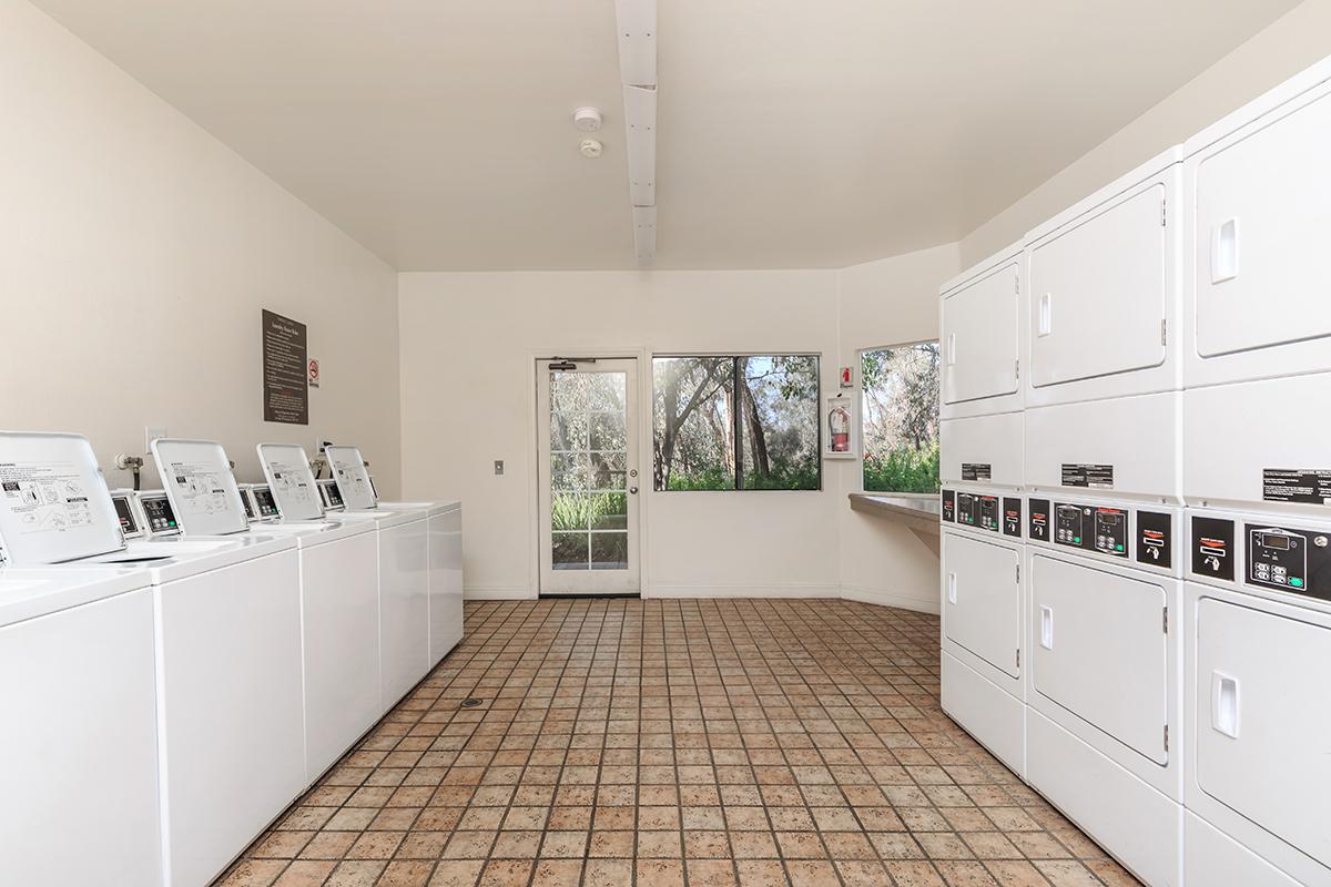 A clean and bright laundry room featuring several white washing machines and dryers lined against the wall. A large window allows natural light to enter, showcasing greenery outside. There is also a countertop space and informational signage on the wall. The floor is tiled in a geometric pattern.