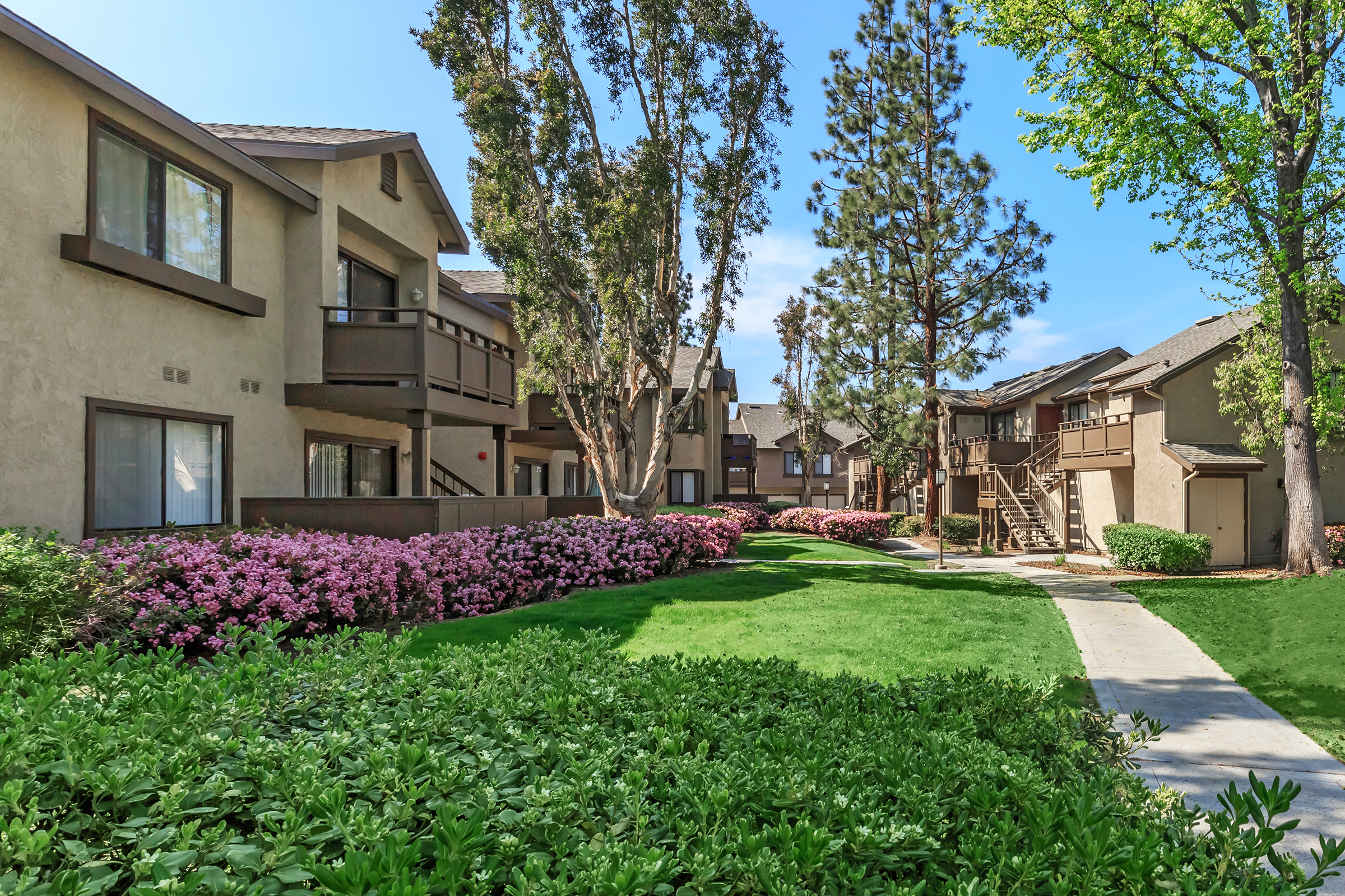 A landscape view of an apartment complex featuring two-story buildings surrounded by lush green grass and colorful flower beds. The scene includes tall trees and a walkway, creating a serene and inviting atmosphere. Bright blue sky complements the vibrant outdoor setting.