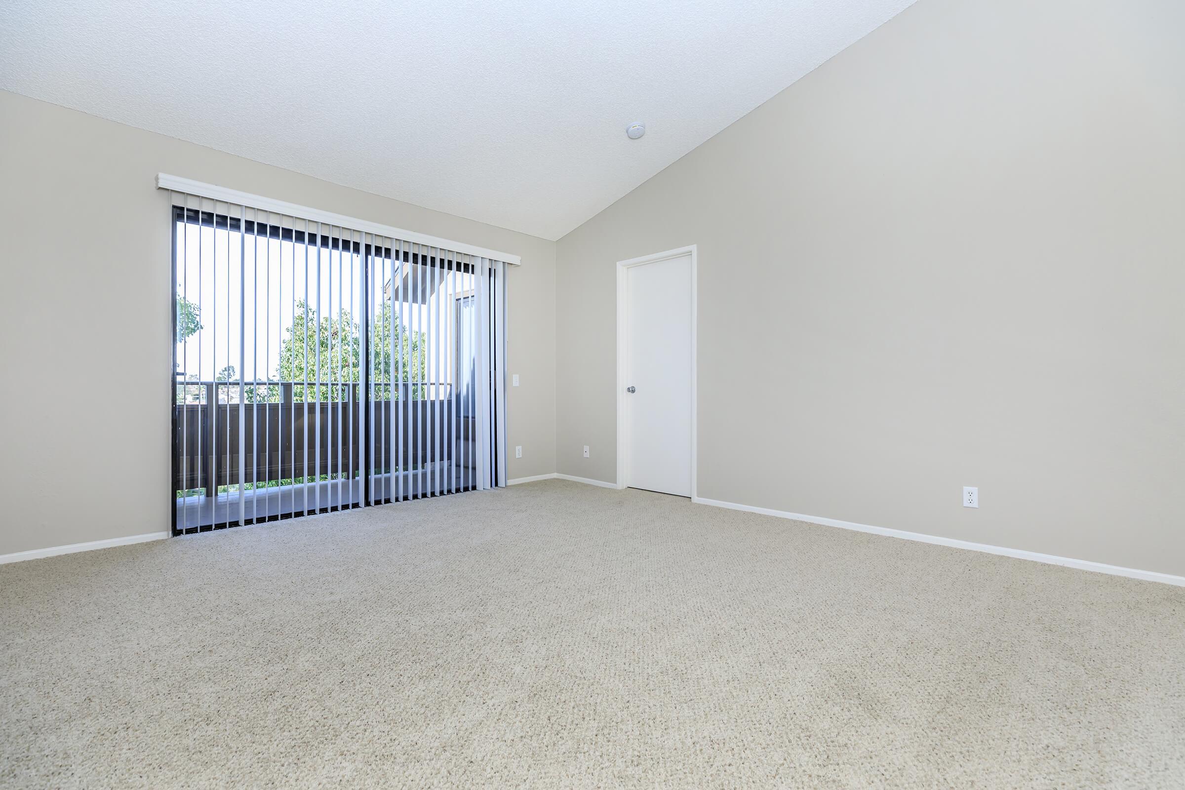 A spacious, empty room with beige walls and light-colored carpet. Large sliding glass doors with vertical blinds lead to a balcony, allowing natural light into the space. A single door on the right side suggests access to another area. The overall clean and minimalistic design offers a blank canvas for decor.