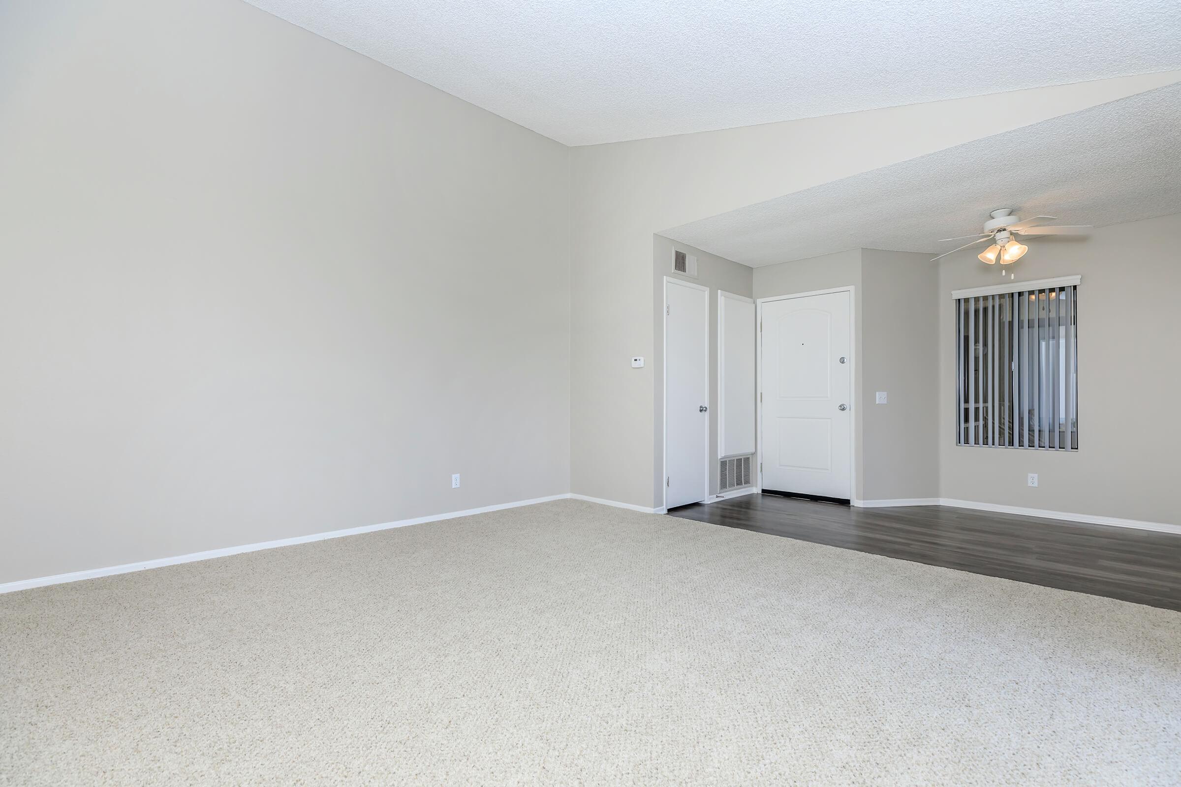 A spacious, empty living room featuring light beige carpeting, beige walls, and a high ceiling. There is a doorway leading to another room, and a door visible on the left side, suggesting access to outside or another area. A ceiling fan is present in the corner of the room. The space is bright and airy.