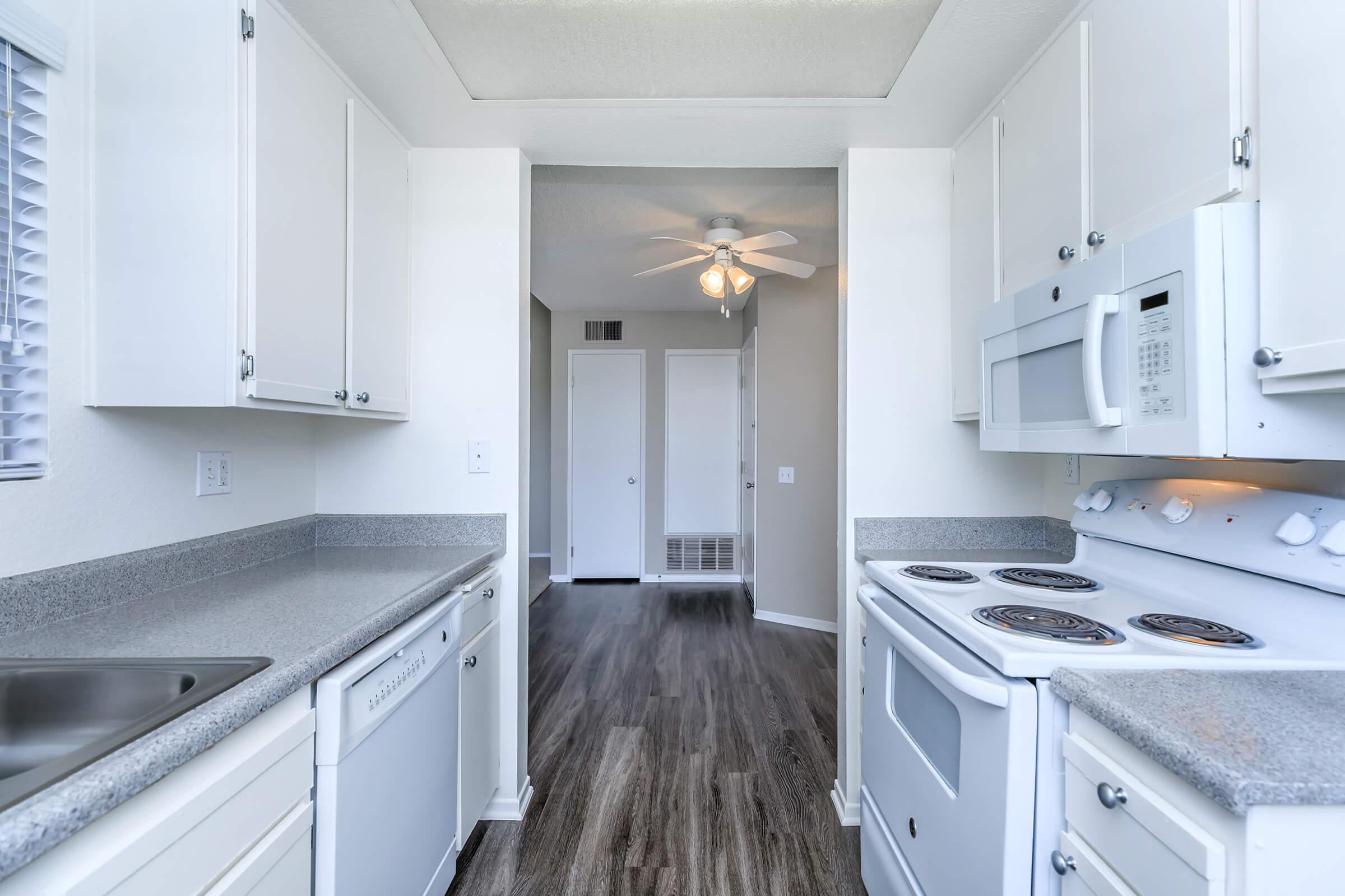 Modern kitchen interior featuring light-colored cabinets, gray countertops, and stainless steel appliances. The space includes a dishwasher, stove, and microwave. A ceiling fan is visible, along with a doorway leading to another room in the background, showcasing an open layout with neutral wall colors and flooring.