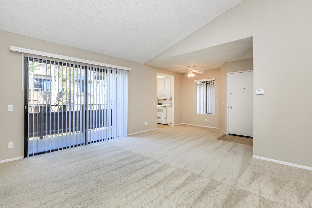 Spacious living area with beige walls and carpeting, featuring large sliding glass doors that lead to a balcony. A ceiling fan adds comfort, while the kitchen can be seen through an open doorway. Natural light fills the room, creating a welcoming atmosphere.