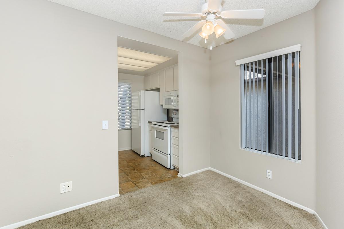 A view of a small, light-colored room with carpeted flooring and a ceiling fan. To one side, there is an open kitchen area featuring a refrigerator, oven, and cabinetry. Large vertical blinds cover a window, allowing natural light to enter the space, which has a neutral color palette.