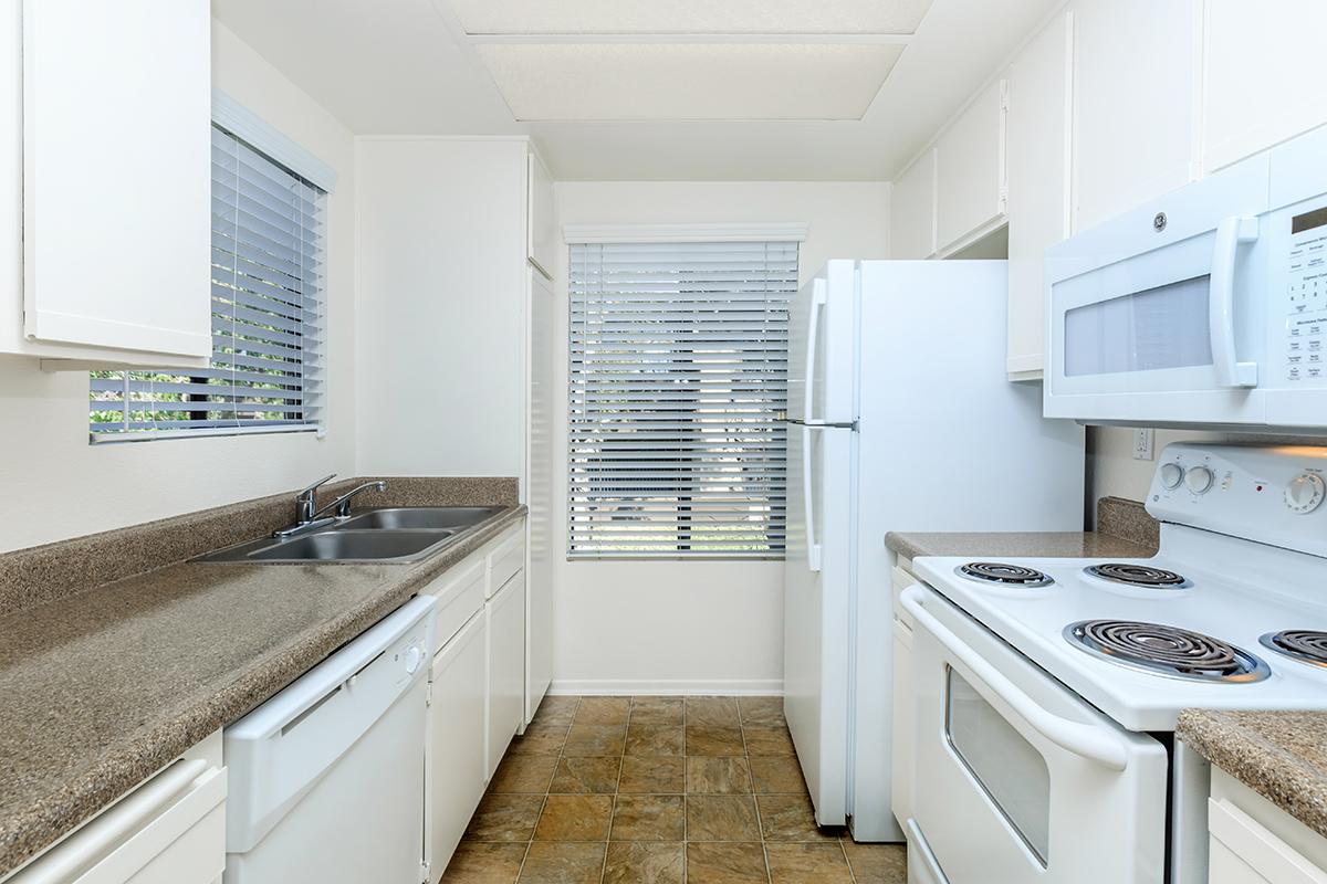 A bright, modern kitchen featuring white cabinetry, a beige countertop, and stainless-steel appliances. The kitchen includes a double sink, a white refrigerator, an electric stove with an oven, and a microwave. Large windows with blinds allow natural light to fill the space.
