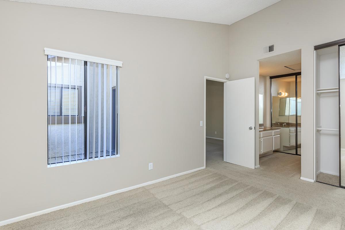 A spacious, empty room with light beige walls and carpet. There is a window with vertical blinds allowing natural light in, and an open door leading to an adjacent bathroom. The room has a high ceiling, creating an airy atmosphere.