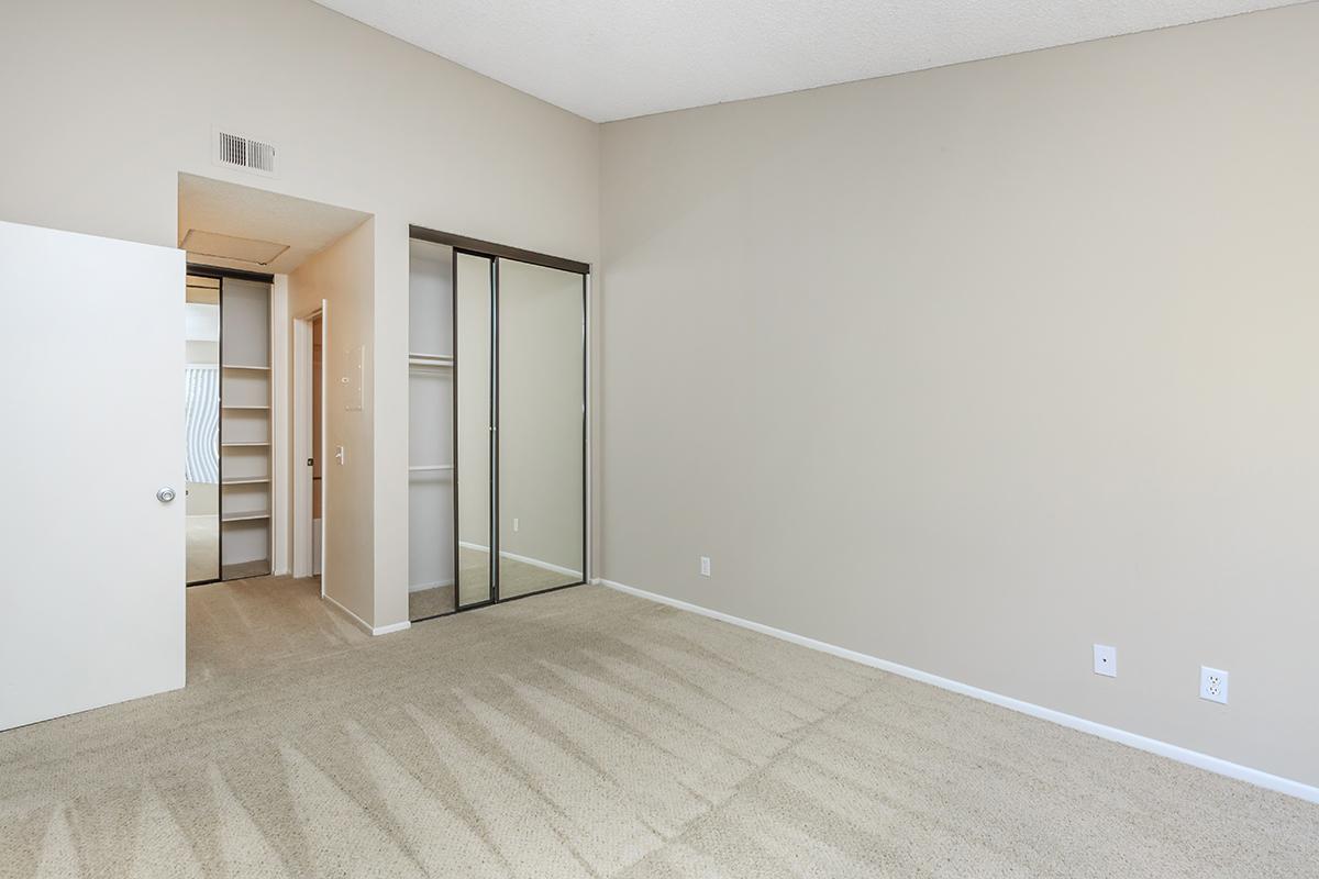 A spacious, empty room with light beige walls and carpeted floor. On one side, there's a door leading to a hallway or closet, and on the opposite side, a large mirrored closet. Natural light creates soft shadows on the carpet, giving the space a bright and open feel.