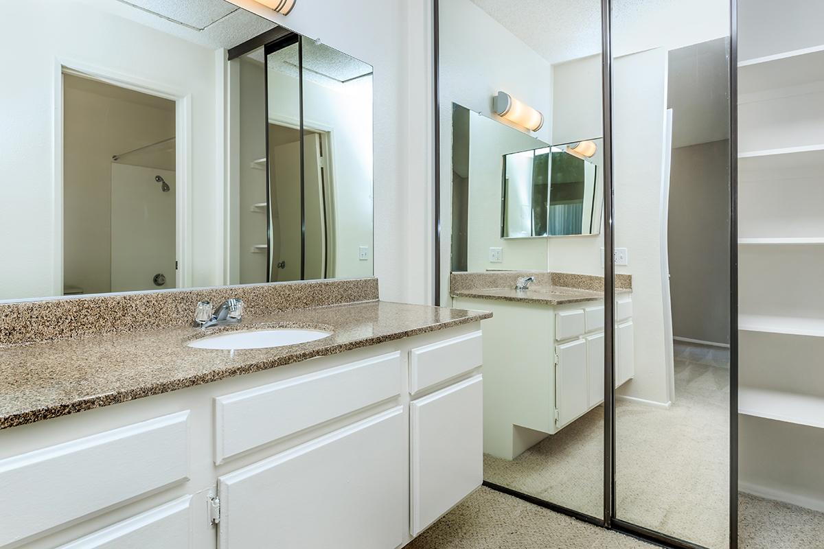 A spacious bathroom featuring a beige granite countertop with a sink, a large mirror, and ample storage cabinets. The walls are painted light, and there is a shower area visible through an open door. The floor is carpeted, creating a cozy atmosphere. Bright lighting fixtures are mounted above the mirror.