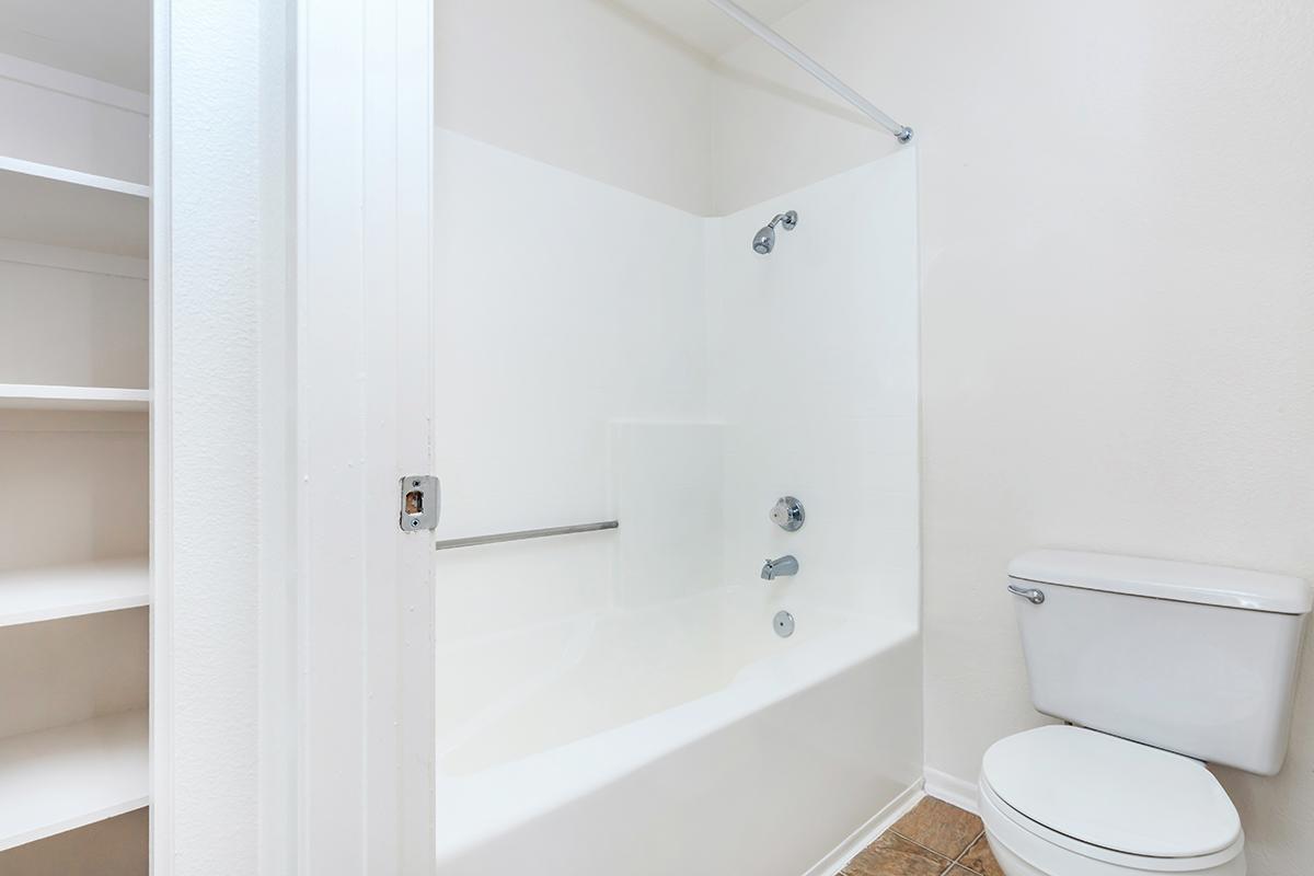 A clean and modern bathroom featuring a bathtub with a shower curtain, a wall-mounted showerhead, and a toilet. White walls and tile flooring create a bright atmosphere. There is a small built-in shelf next to the bathtub for storage.