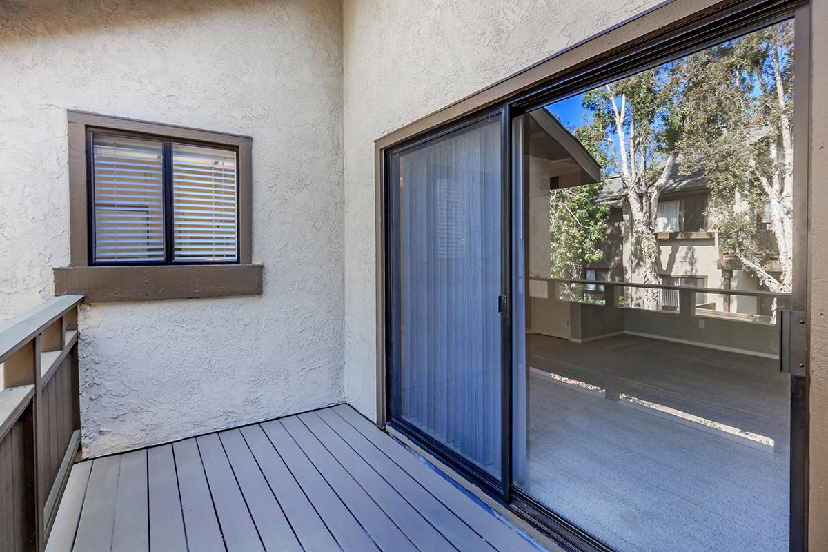 A small balcony area with a sliding glass door leading to an indoor space. The floor is wooden with light gray planks, and there is a window with horizontal blinds on the adjacent wall. Surrounding greenery is visible through the glass, providing a view of the outdoors.