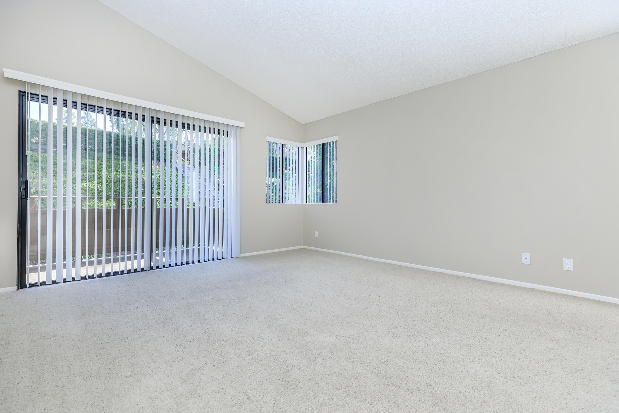 Spacious, empty room with light-colored carpet and beige walls. Large windows with vertical blinds allow natural light to enter, complemented by a sliding glass door leading to a balcony. The room features a high ceiling, creating an open and airy atmosphere. Ideal for customization and decor.
