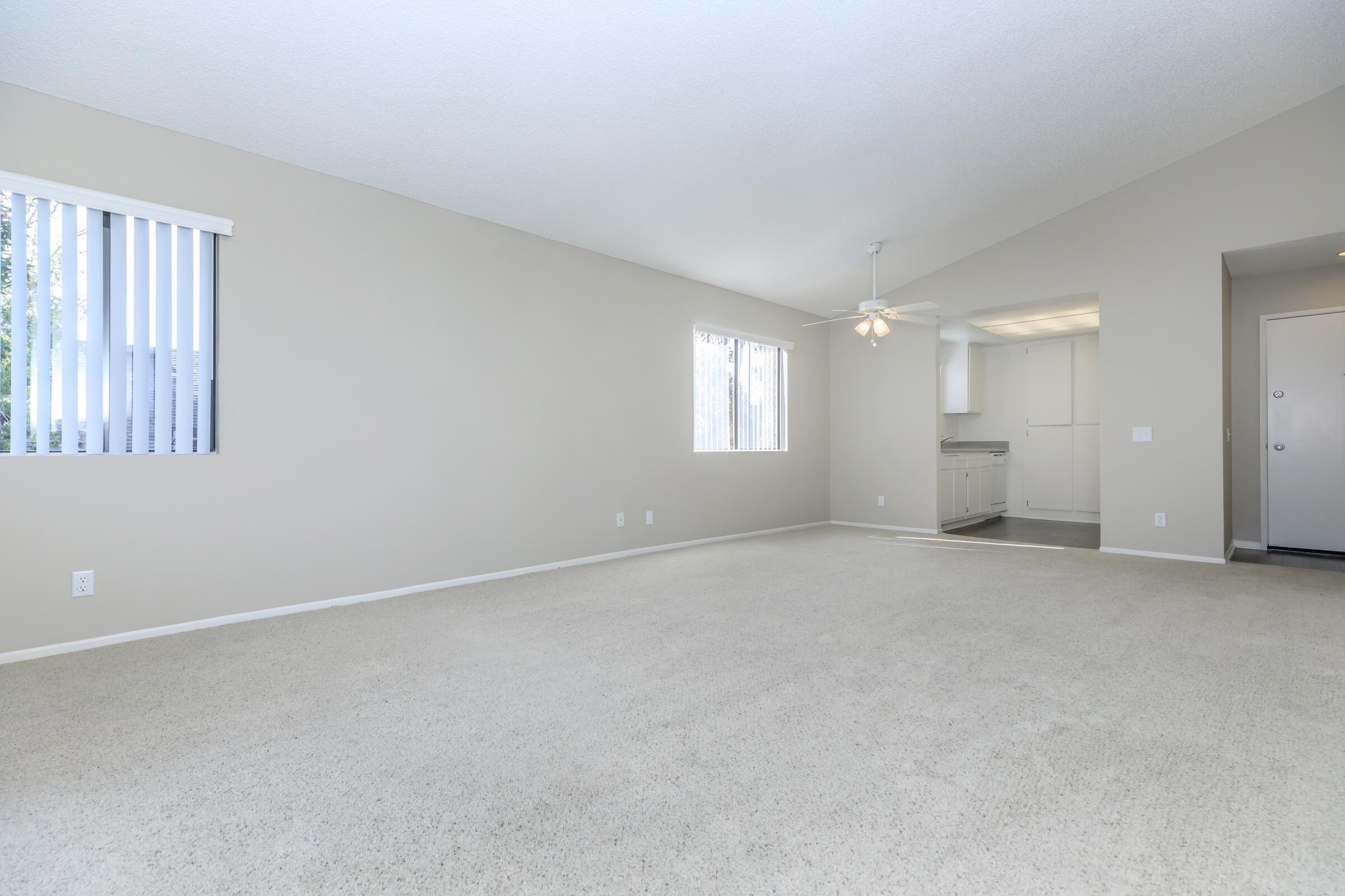 Spacious, empty living area with light beige carpet and neutral-colored walls. A window with vertical blinds allows natural light, and a ceiling fan is visible. In the background, there is a glimpse of a kitchen area and another doorway.